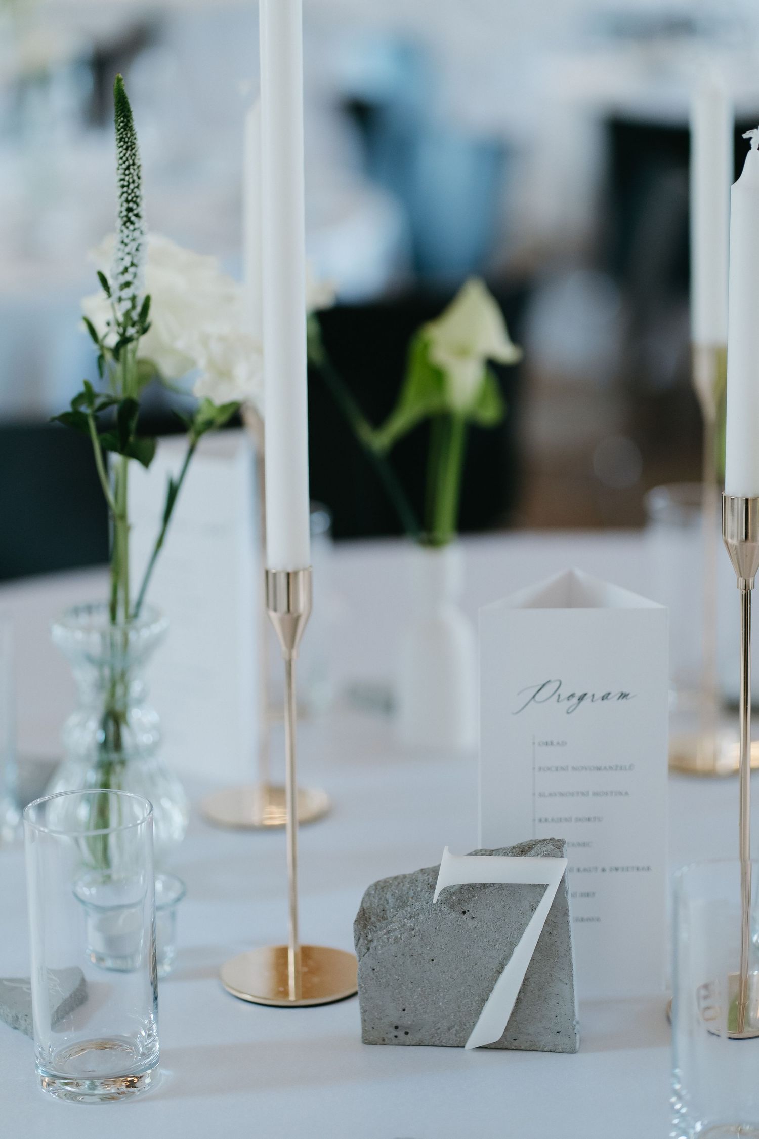 Elegant wedding table with white taper candles in gold holders, a concrete number seven card, bud vases with flowers, and a printed program.