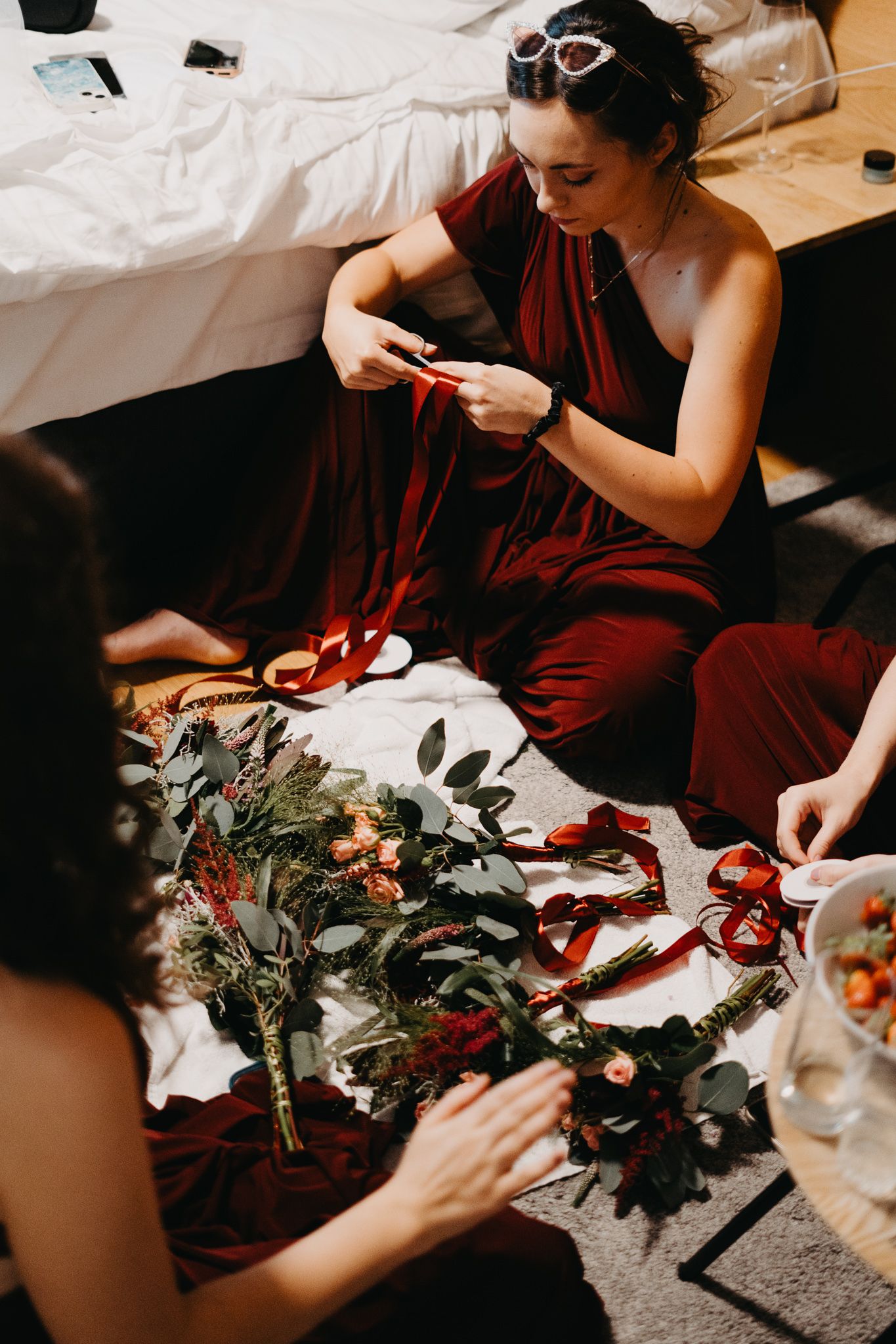 Woman in burgundy dress arranges flowers and ribbons on white surface while crafting holiday decorations.