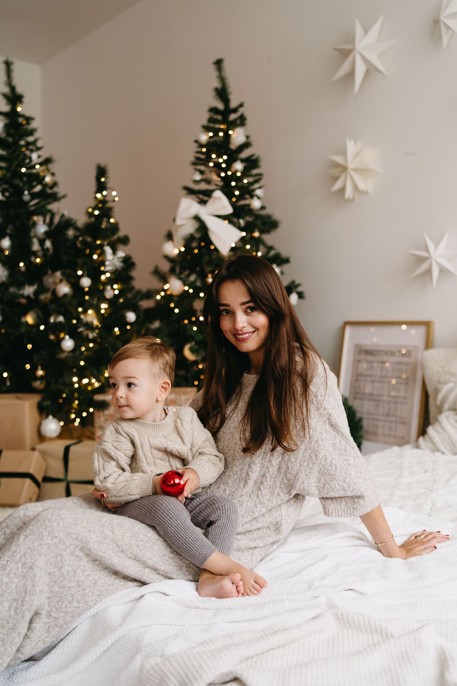 Mother and child in white sweaters share tender Christmas morning moments on bed with decorated trees and stars.