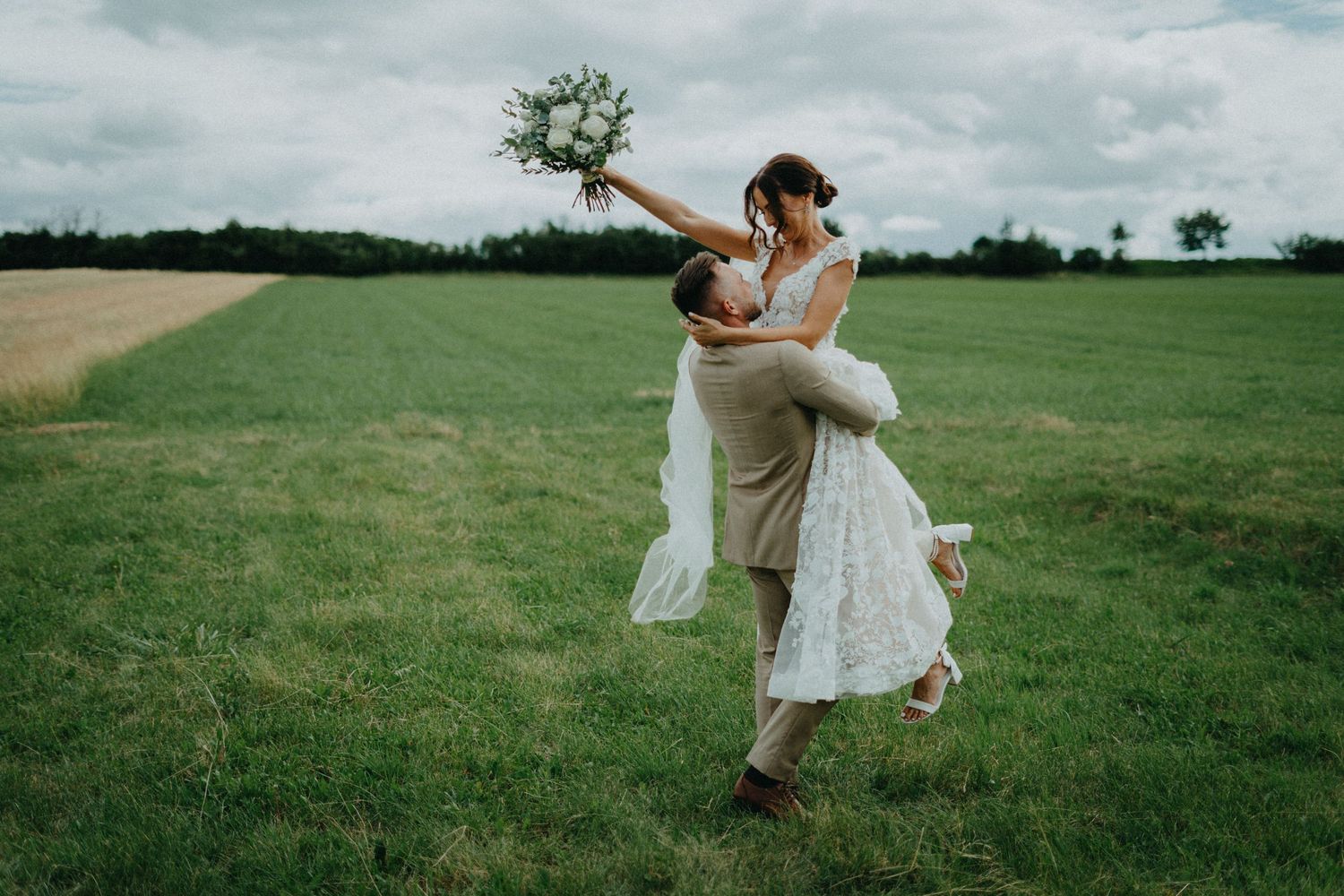 A couple in light summer attire embraces in a vast green meadow under cloudy skies.