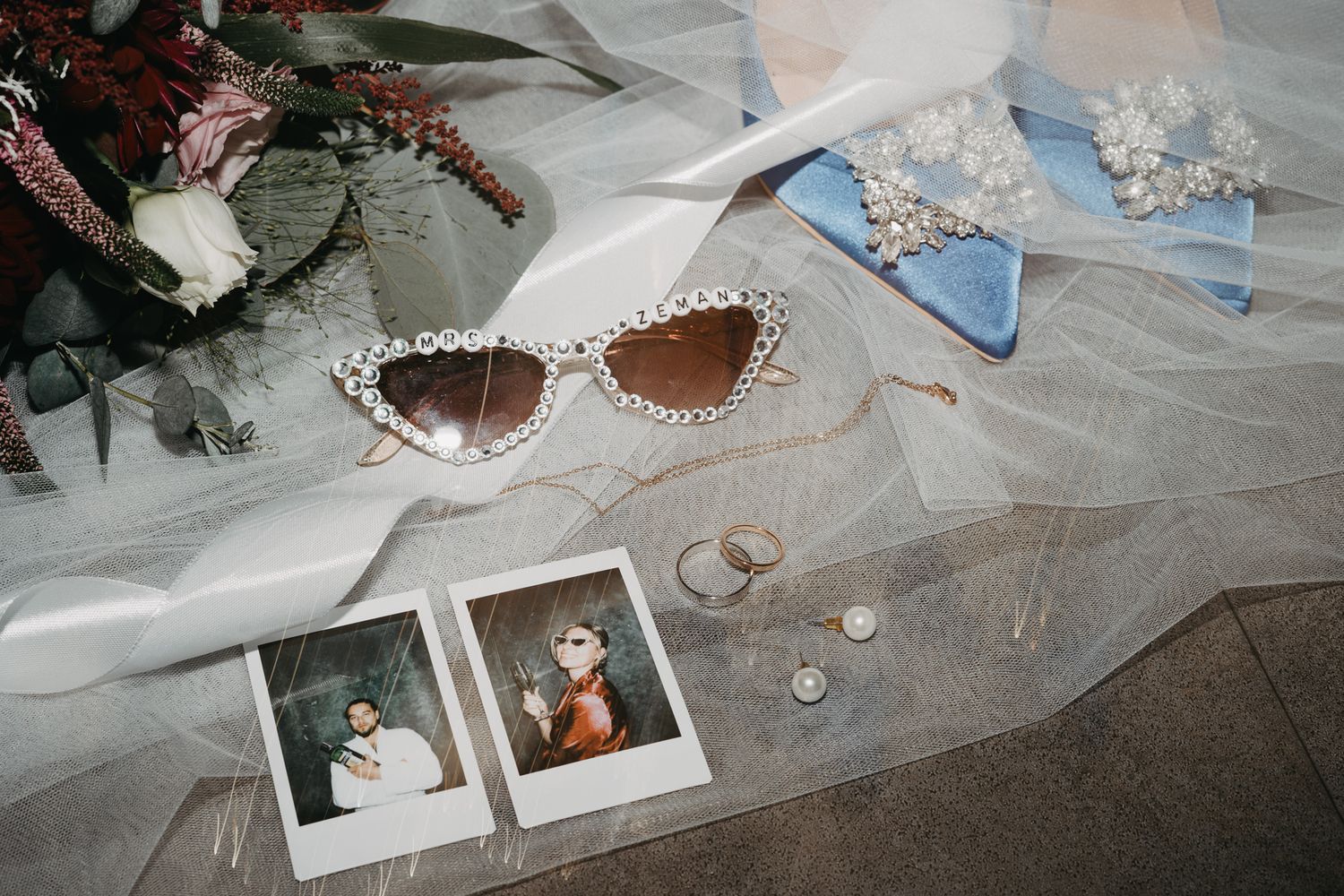 Wedding accessories including retro sunglasses, polaroid photos, jewelry and lace veil arranged on light fabric.