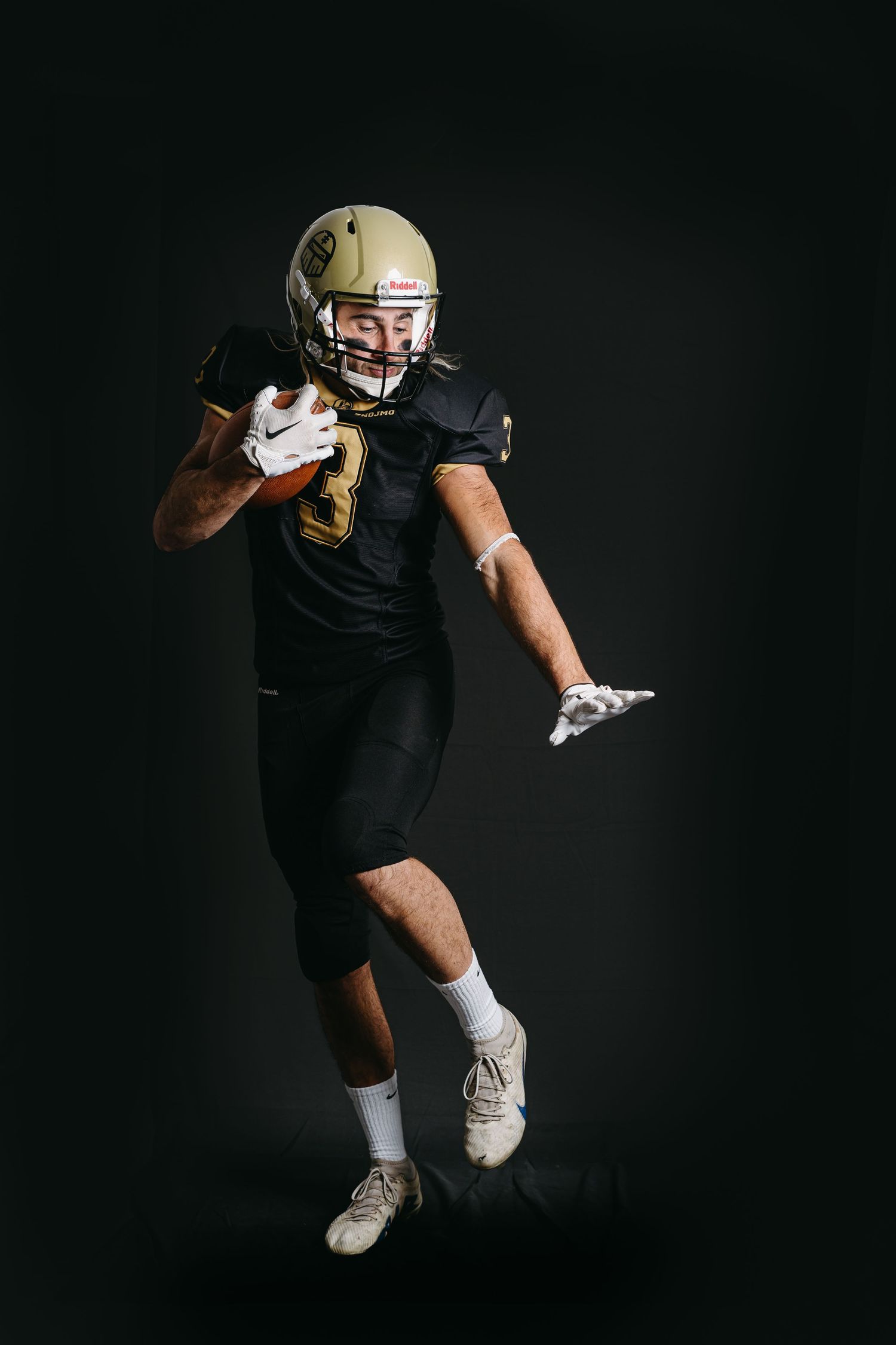 Football player in black and gold uniform sprints and prepares to make a play against dark backdrop.