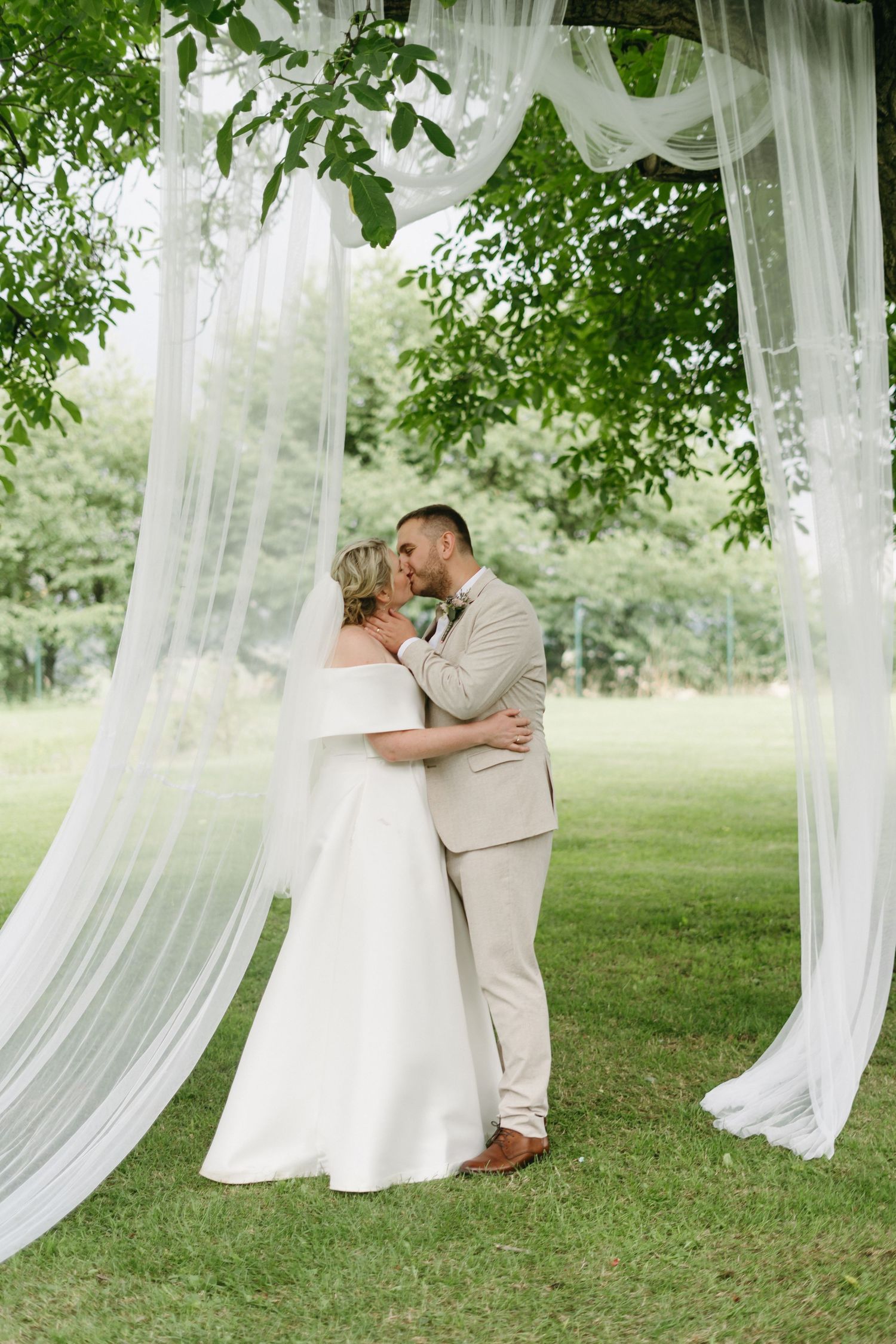 A romantic outdoor wedding ceremony under a flowing white fabric archway amongst green trees and grass.