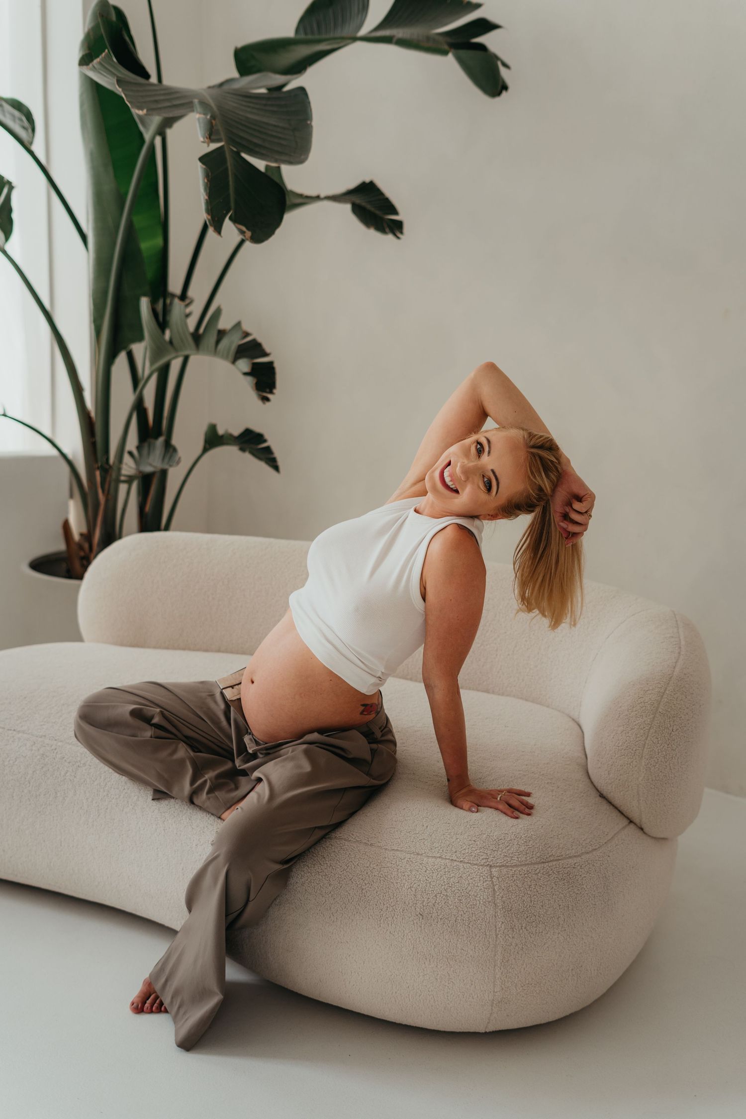 Lifestyle portrait on a cream curved sofa with indoor plants, featuring casual white top and brown pants in natural light setting.