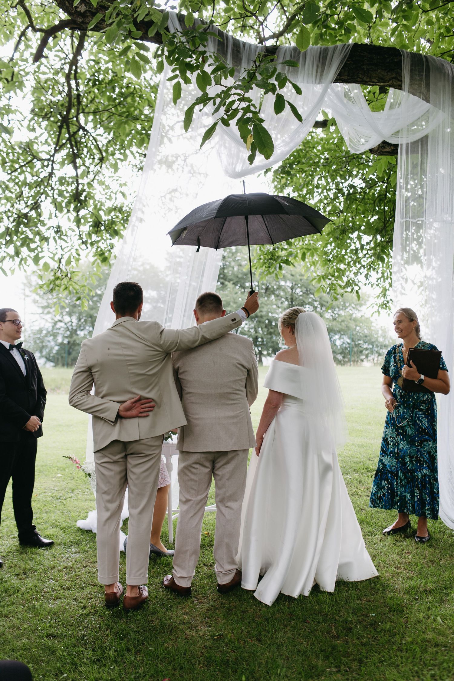 Wedding party members in beige and teal attire pose against a backdrop of white curtains and green trees in the rain.