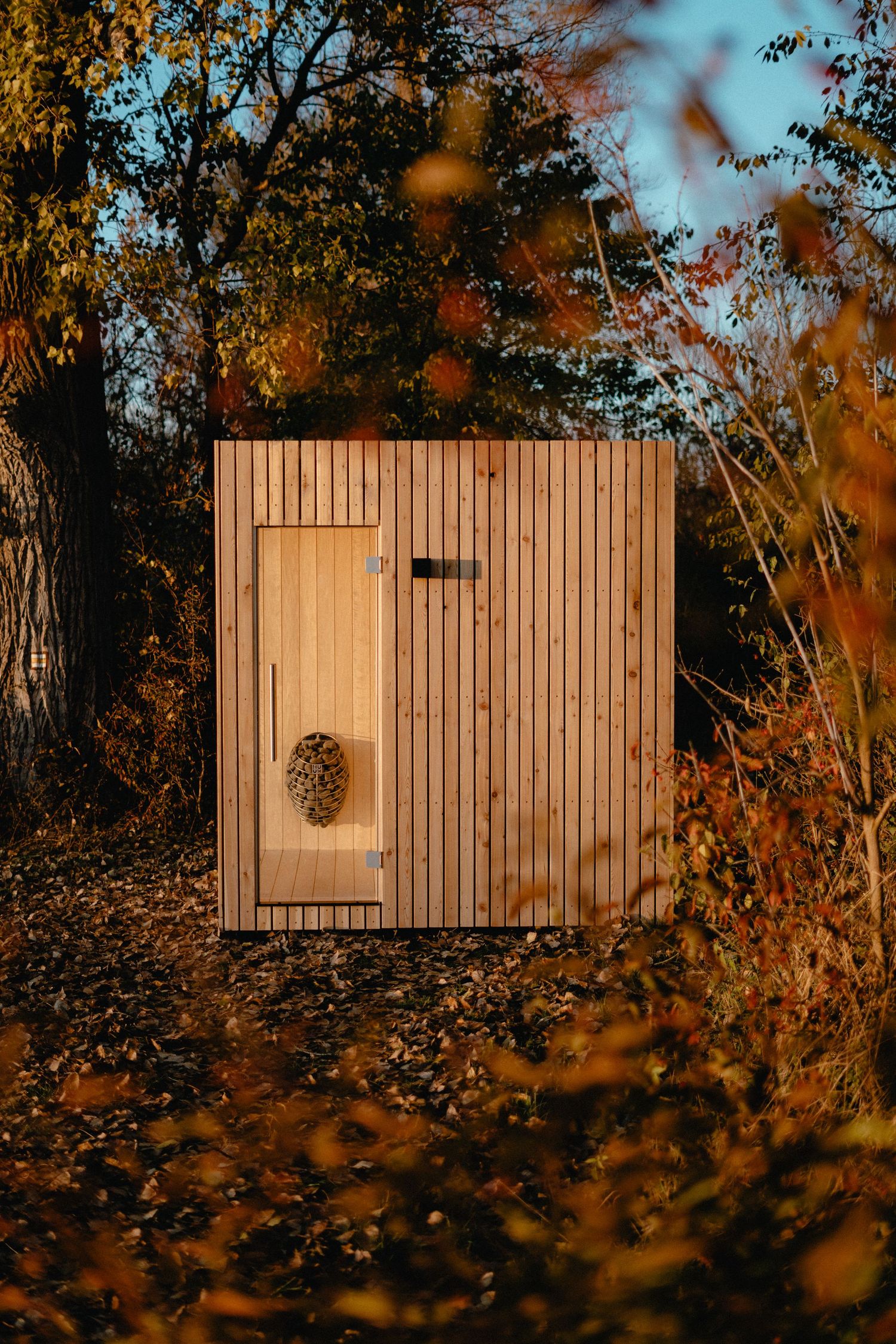 A wooden structure with vertical planks is photographed from different angles against autumn foliage at sunset.