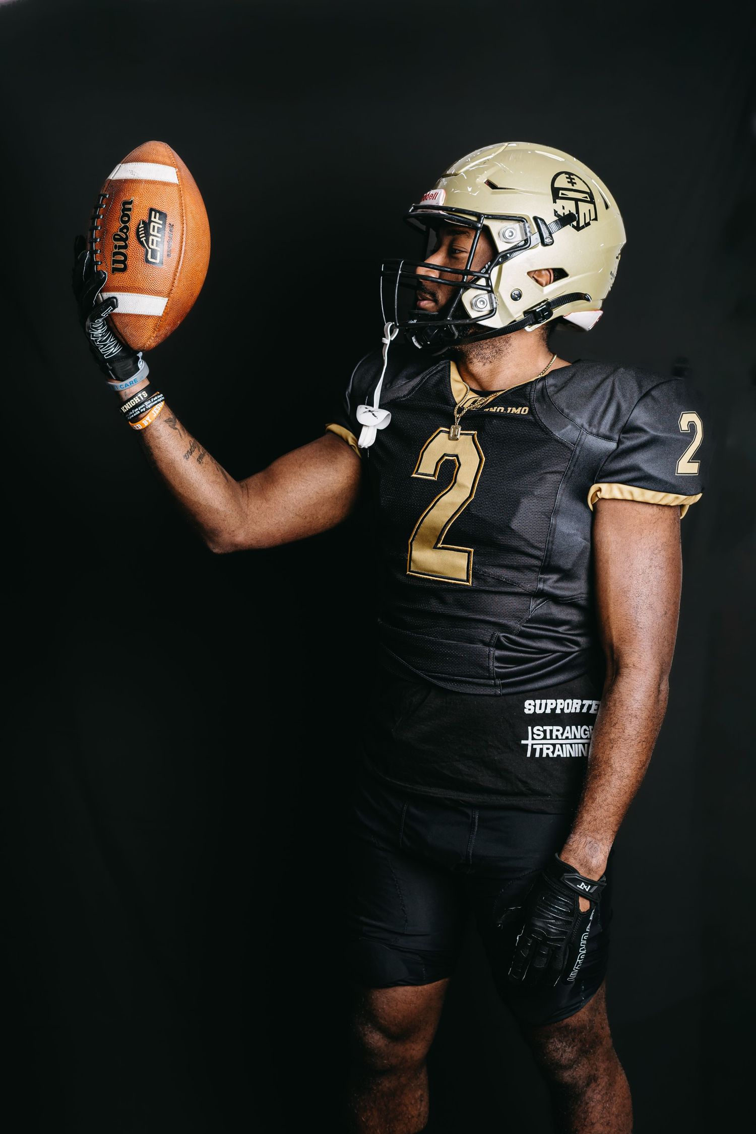 Football player in black and gold uniform showcasing a football against dark backdrop.