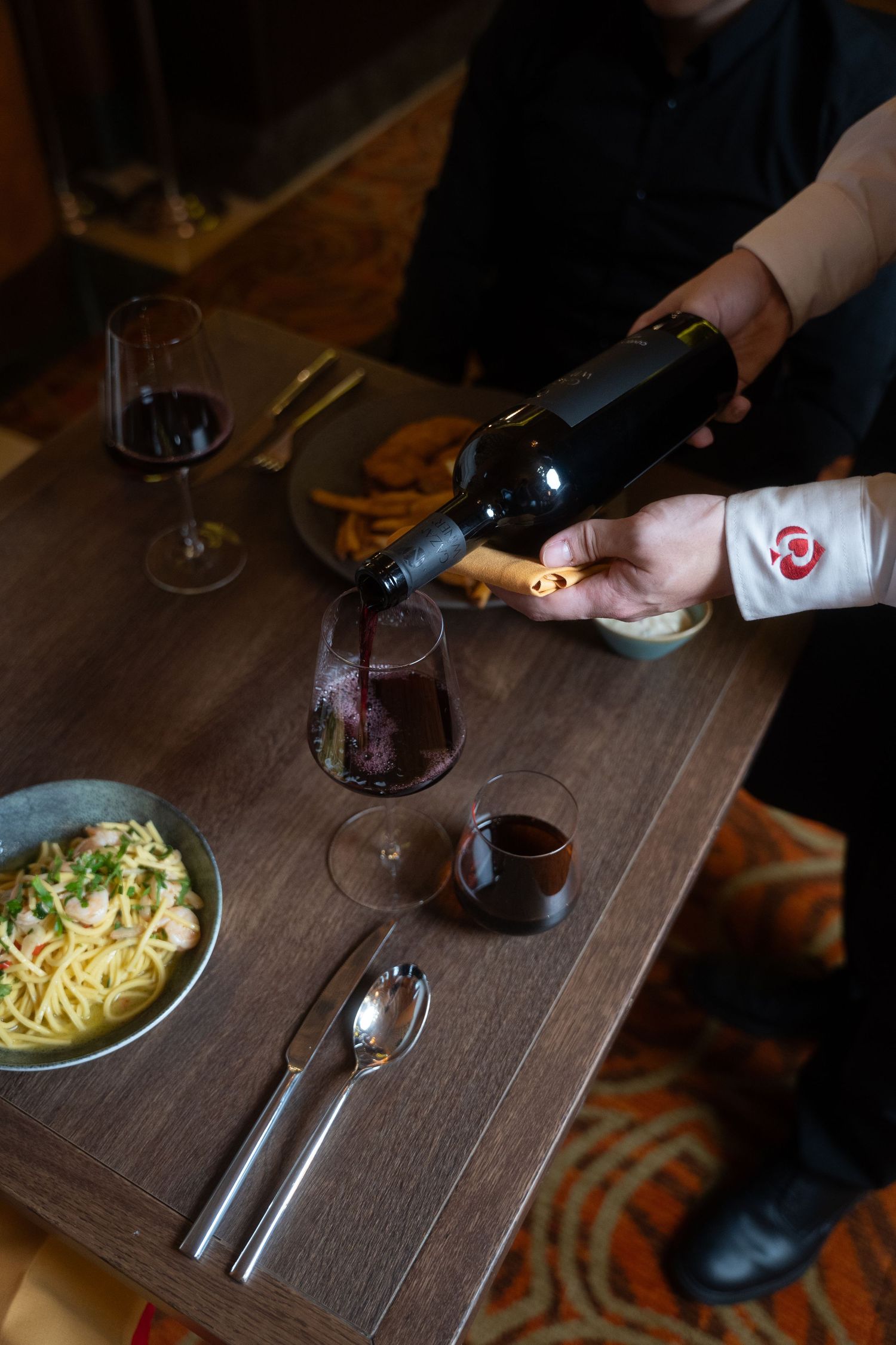 Red wine being poured at dinner table with pasta dish and place settings on dark wooden surface.