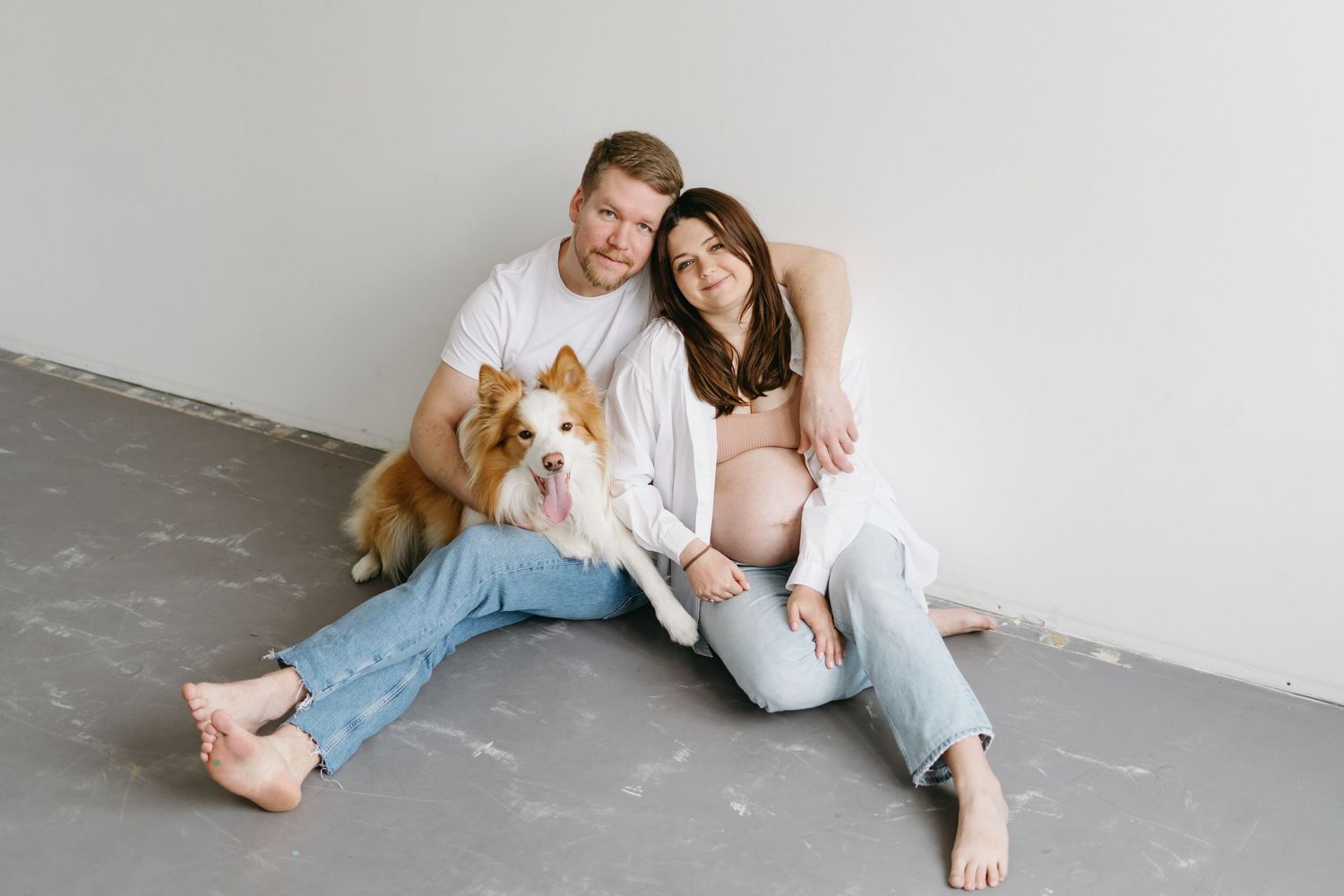 A casual maternity photo featuring a couple and their dog sitting on a concrete floor.