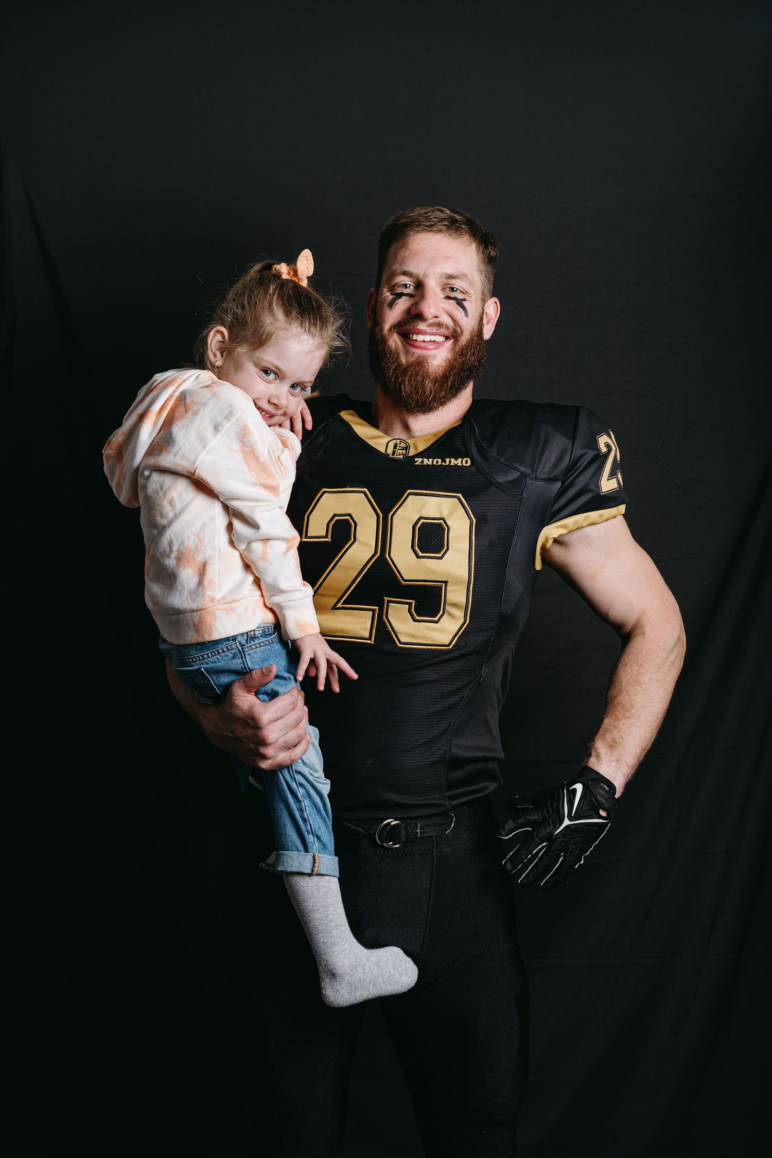 Black-uniformed football player wears number 29 jersey while interacting with a small child against dark background.
