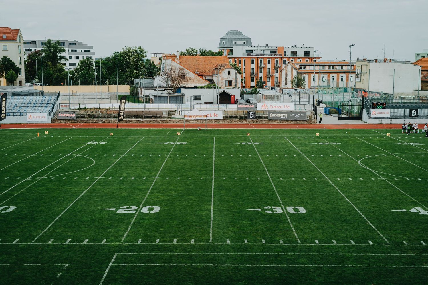 Green artificial turf football field with yard lines marked showing the 30-40 yard area with buildings visible in background.