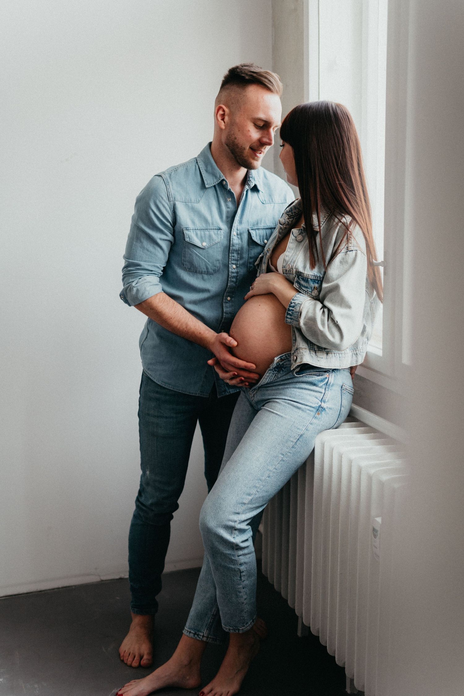 A couple poses by a window during their maternity session wearing casual denim outfits.