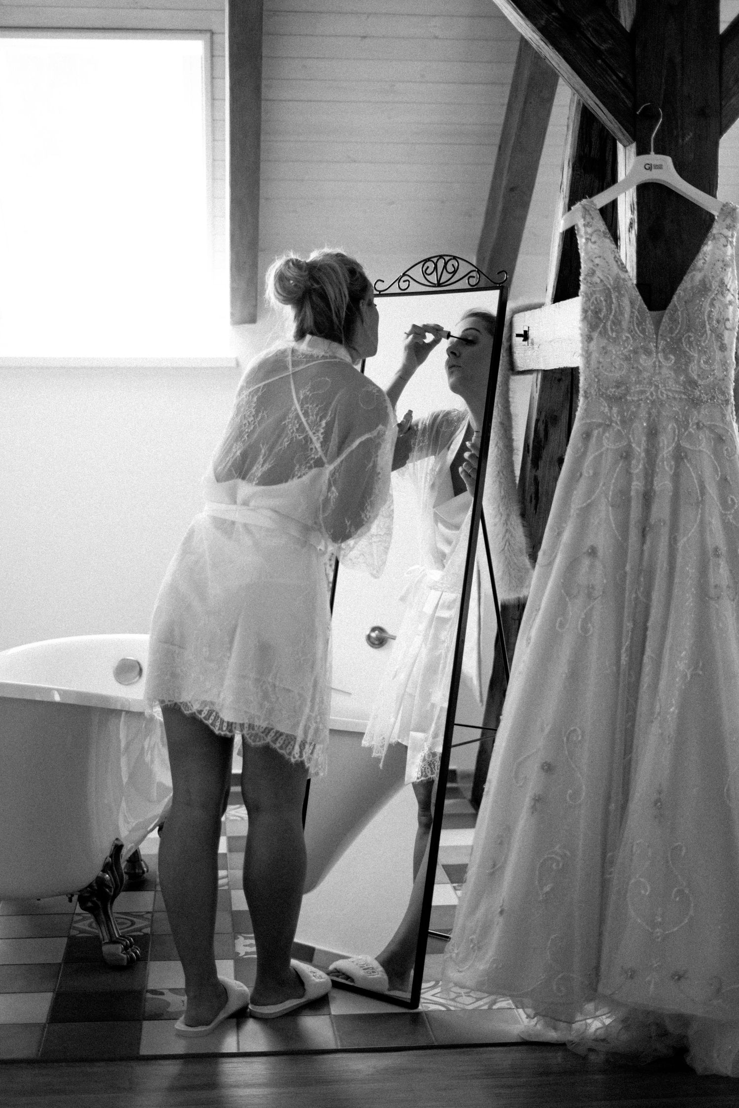 Black and white image of someone getting ready near a wedding dress hanging in a room.