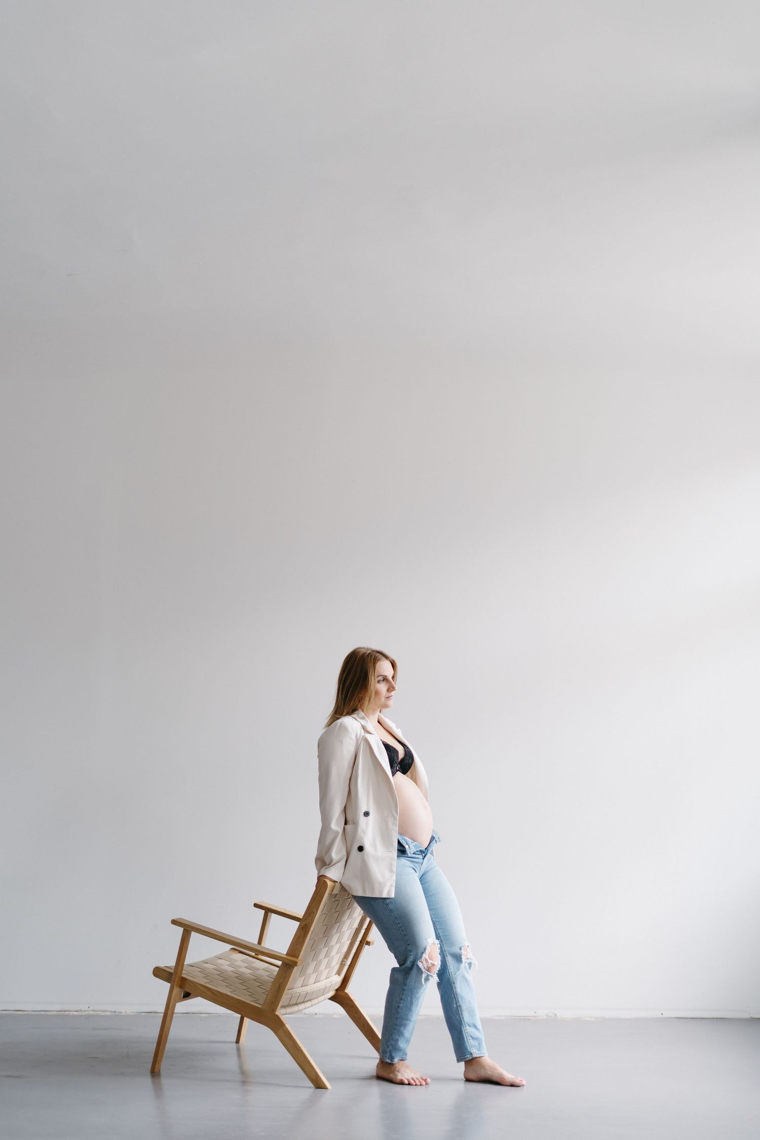 Minimalist maternity session with wooden chair against white wall shows quiet contemplation.