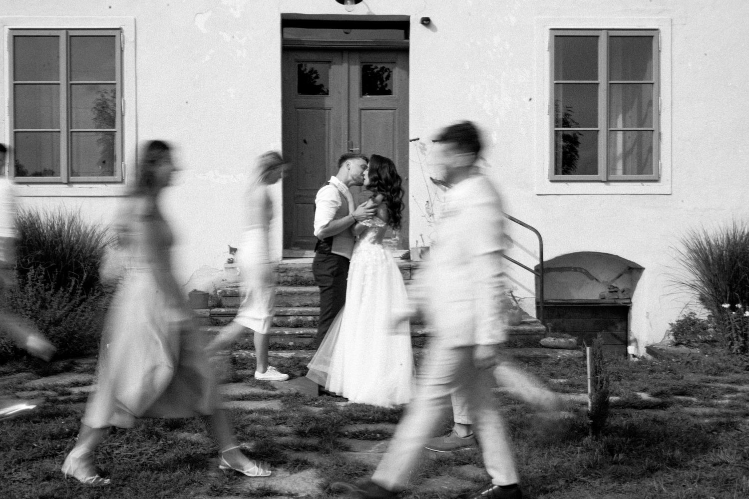 Motion blurred figures in white clothing move across a building facade with windows in a black and white photograph.