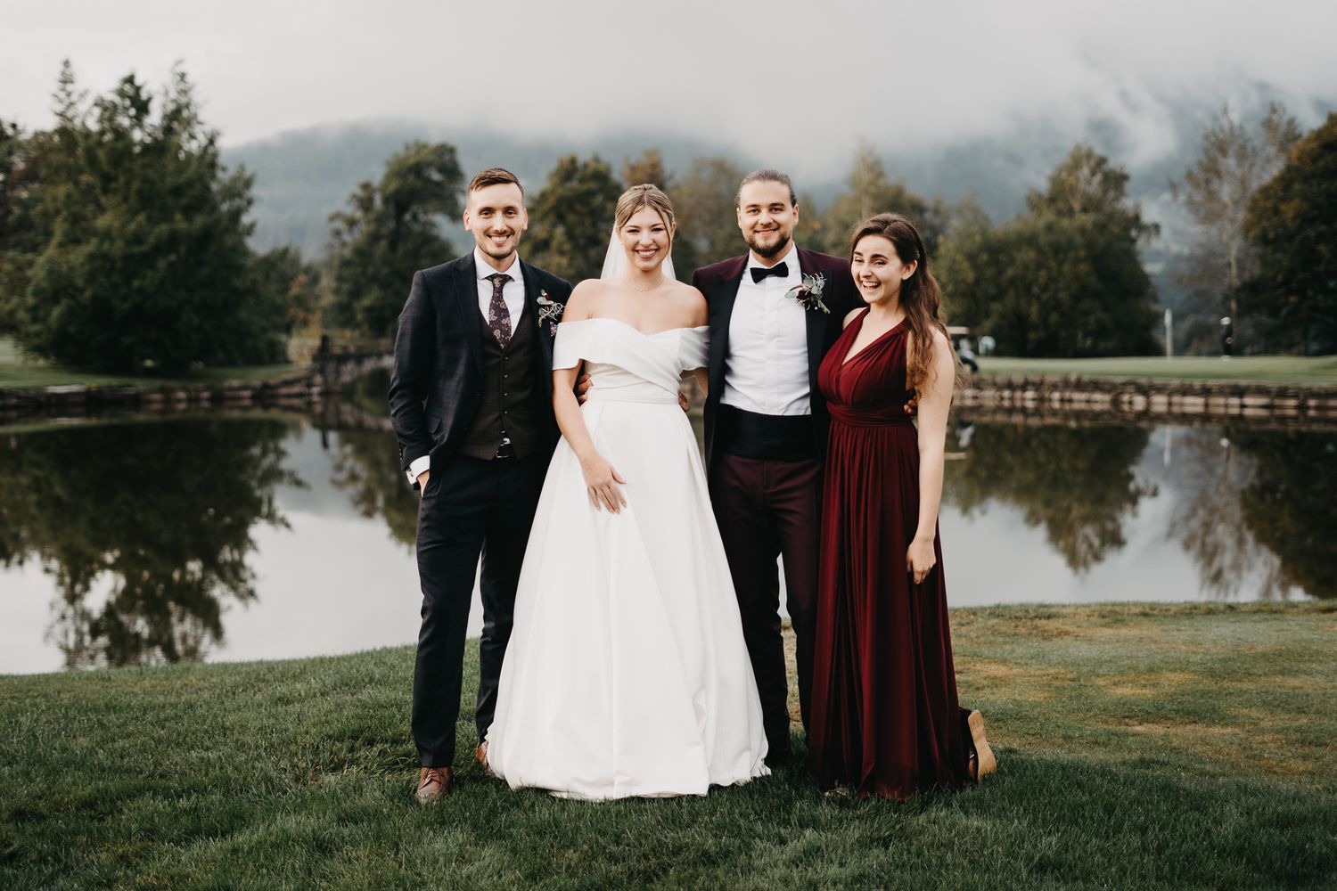 Four people in formal attire pose together by a reflective lake with mountains in the background at a wedding.