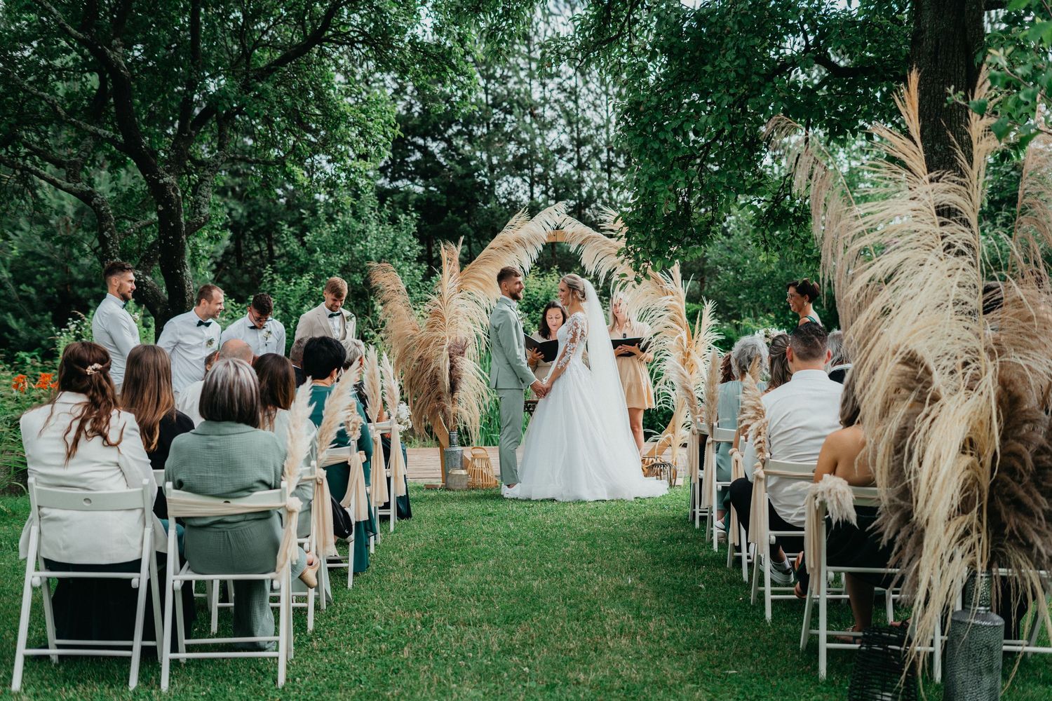 Outdoor wedding ceremony on green lawn with white chairs and pampas grass decorations along the aisle.