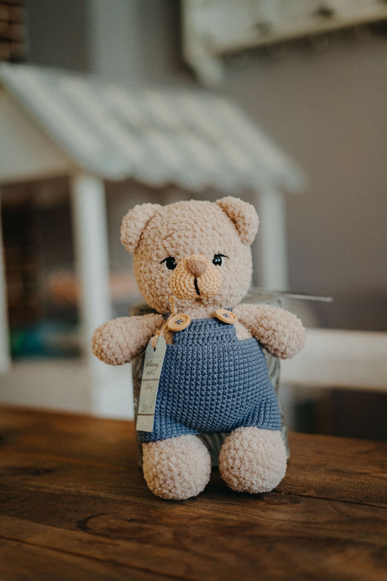 Vintage teddy bear wearing blue knit overalls sits on wooden table near dollhouse in soft focus lighting.