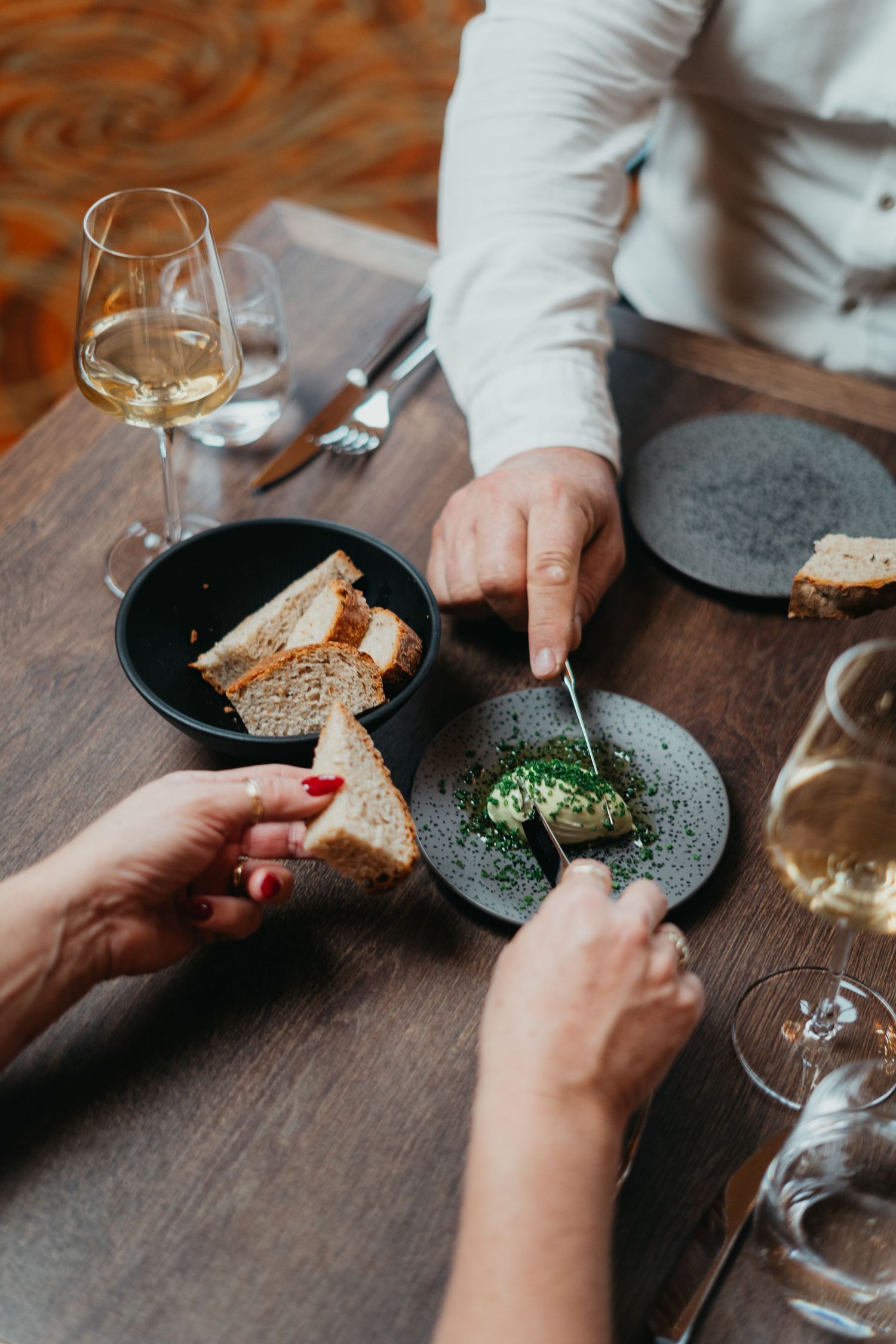 A sequence of hands sharing food and dining at a restaurant table with wine glasses and dark plates.