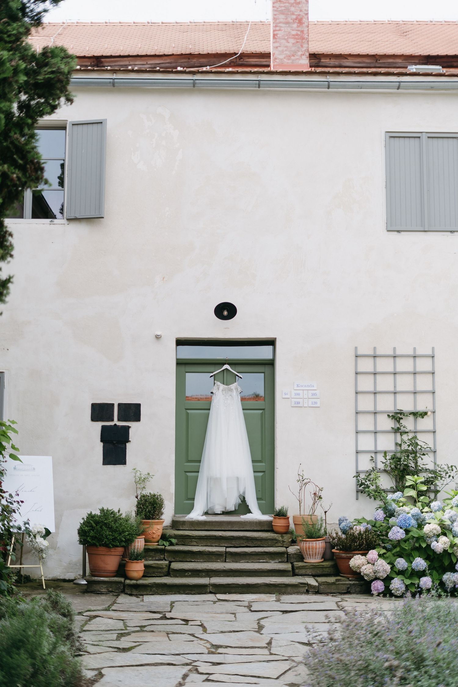 Wedding gown displayed on white exterior of historic building with green shutters and stone pathway surrounded by flowers.