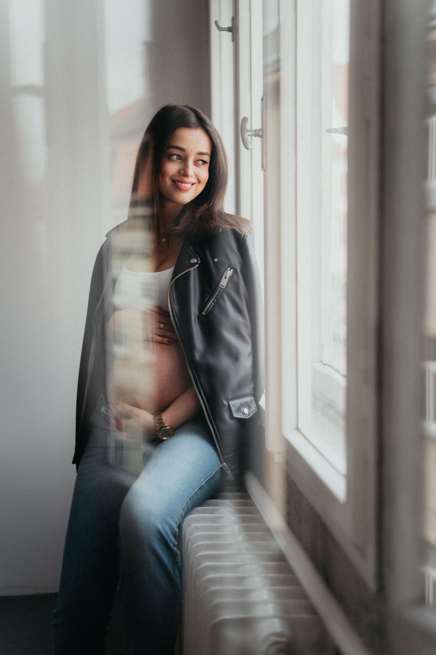 Maternity portrait by window with woman wearing leather jacket in natural light setting.