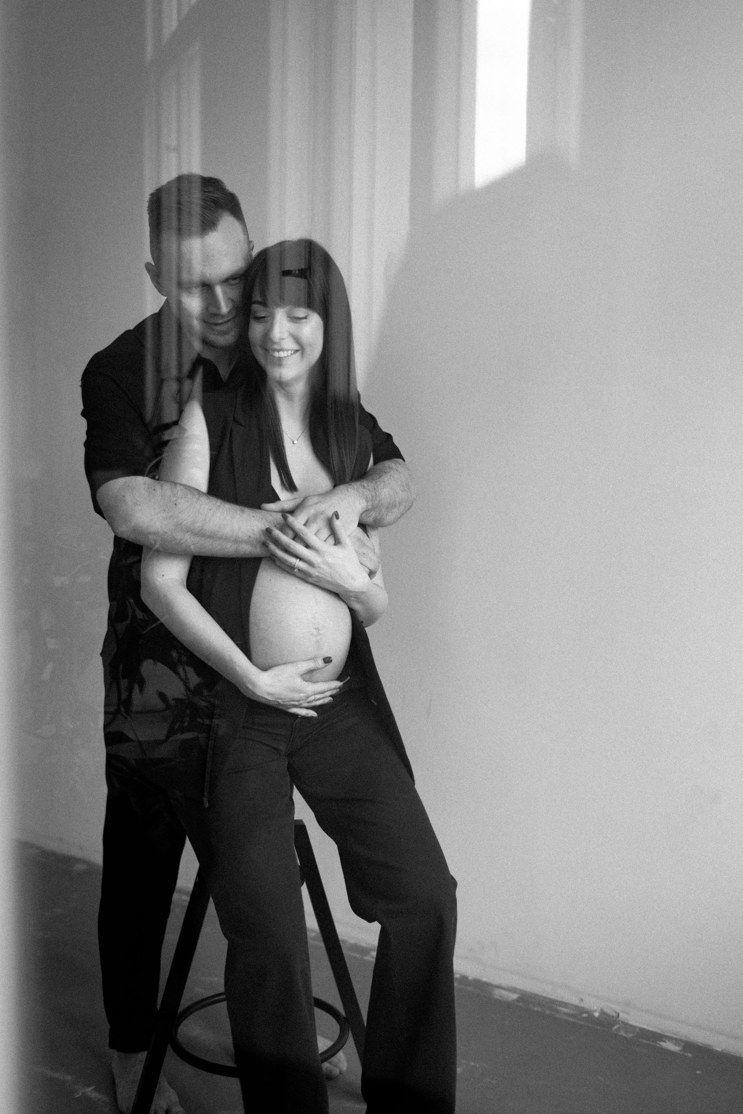 A tender black and white maternity photo of a couple embracing against a wall with soft window light.