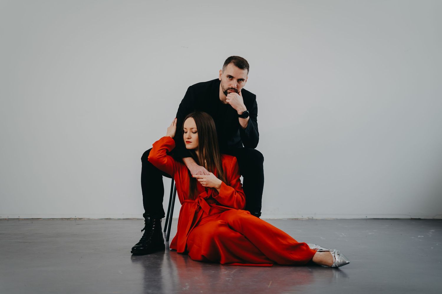 Couple in stylish outfits posing together on grey floor during indoor photoshoot.