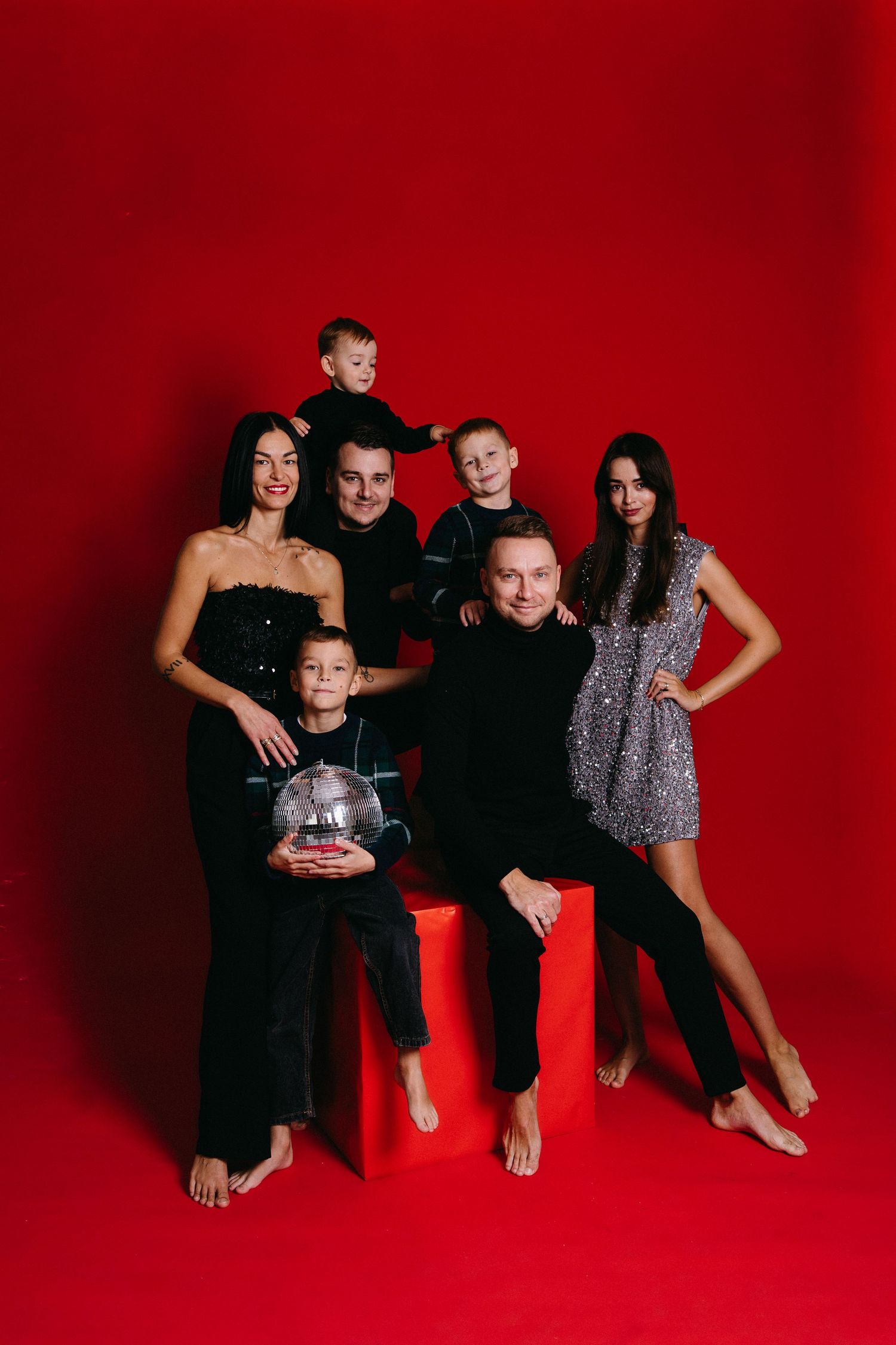 Family group poses together against vibrant red backdrop in coordinated black outfits for studio portraits.