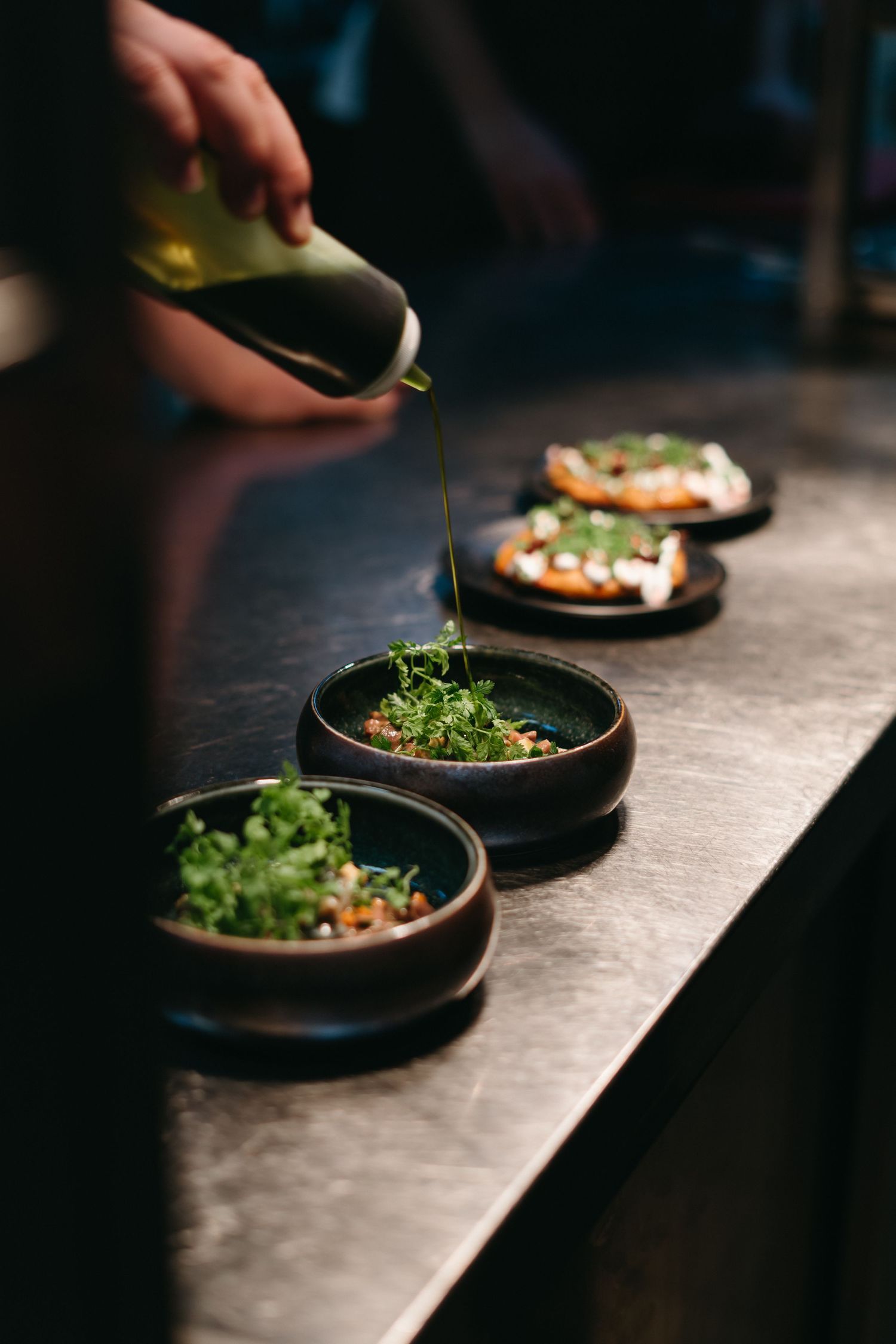 Chef drizzling oil over fresh green salads in dark ceramic bowls on a restaurant counter.
