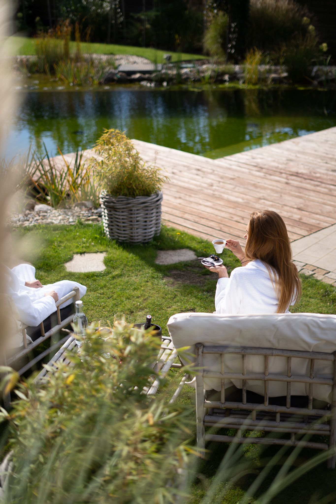 Person relaxing on outdoor wooden deck furniture by a tranquil pond on a sunny day.