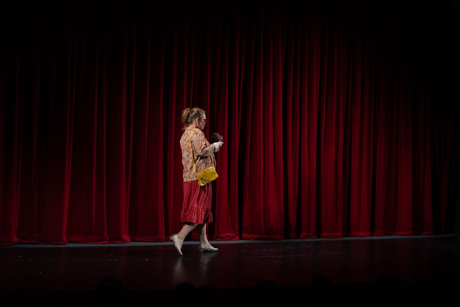 Solo performer dances gracefully in a black dress against dramatic red stage curtains under theatrical lighting.
