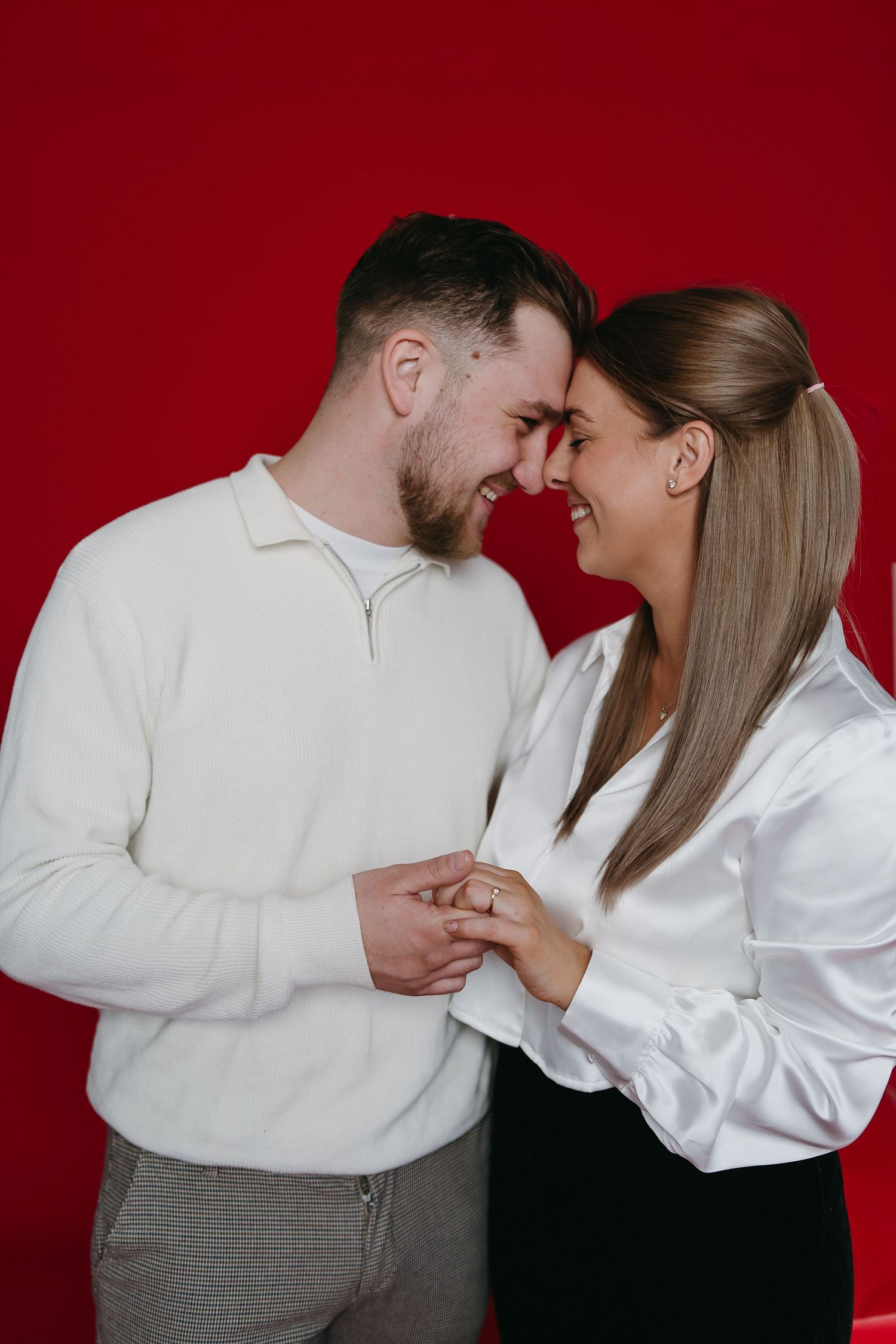 A smiling couple in white tops hold hands and touch foreheads against a bold red background.