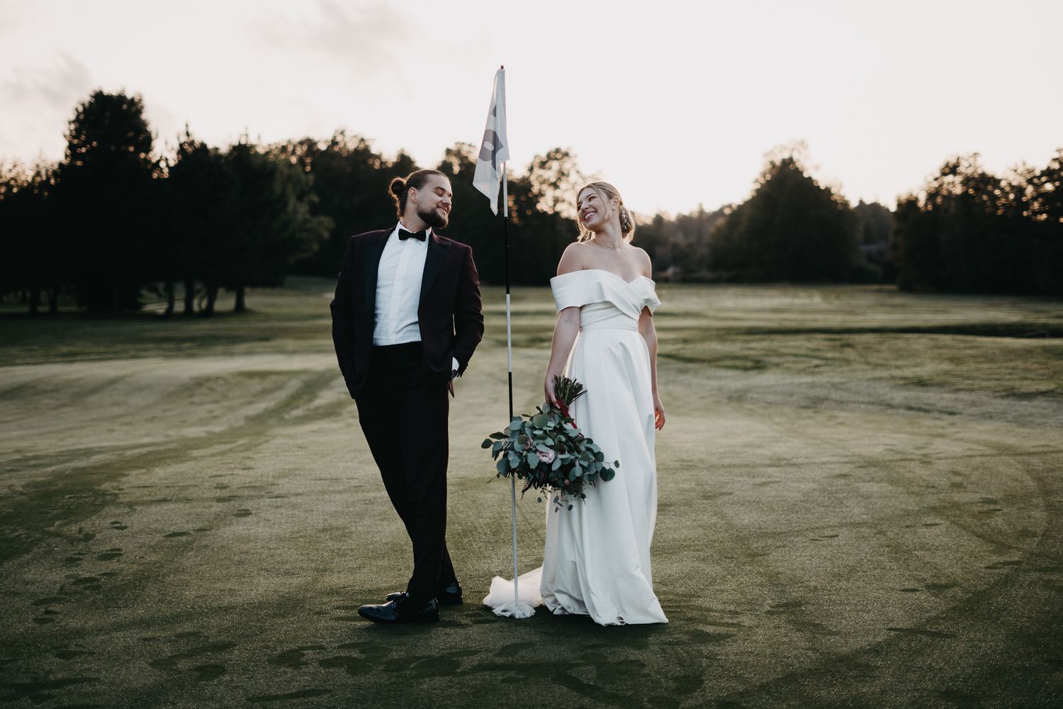 A couple stands together on a golf course at sunset in formal wedding attire holding a blue floral bouquet.