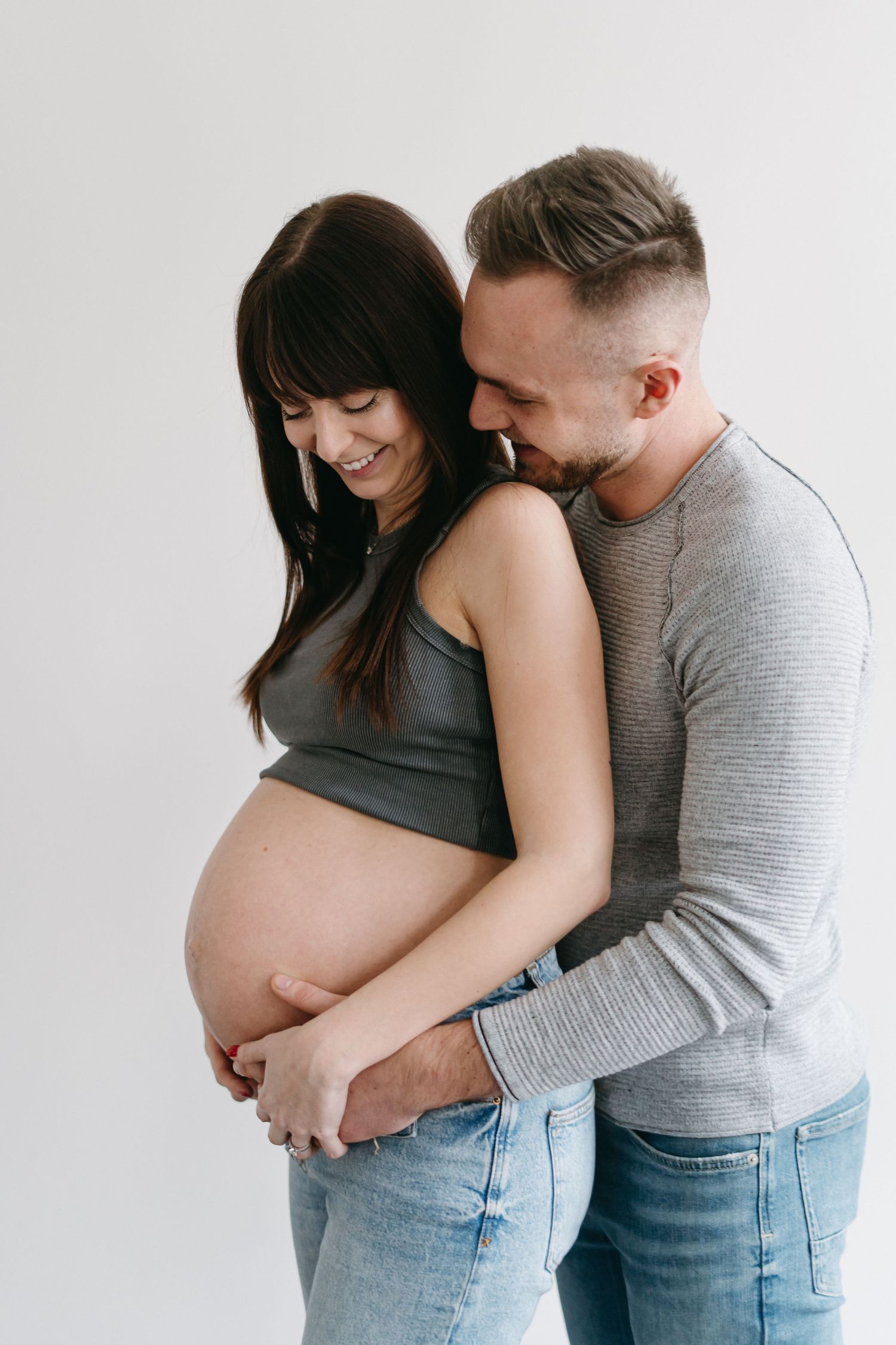 Expecting couple shares an intimate moment as they embrace the pregnant belly against a white backdrop.