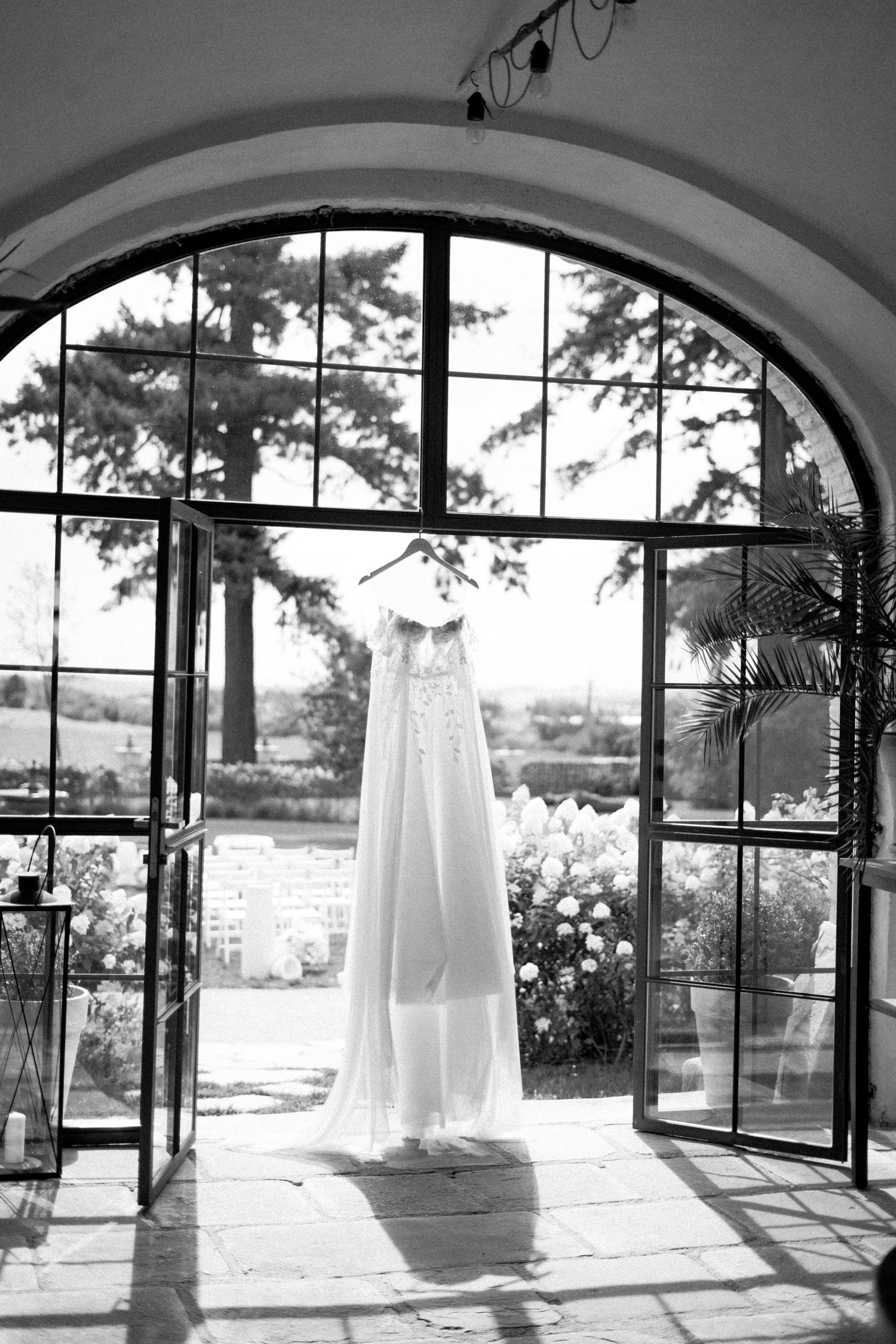 A white wedding dress hangs elegantly in an arched doorway with scenic garden views in black and white.