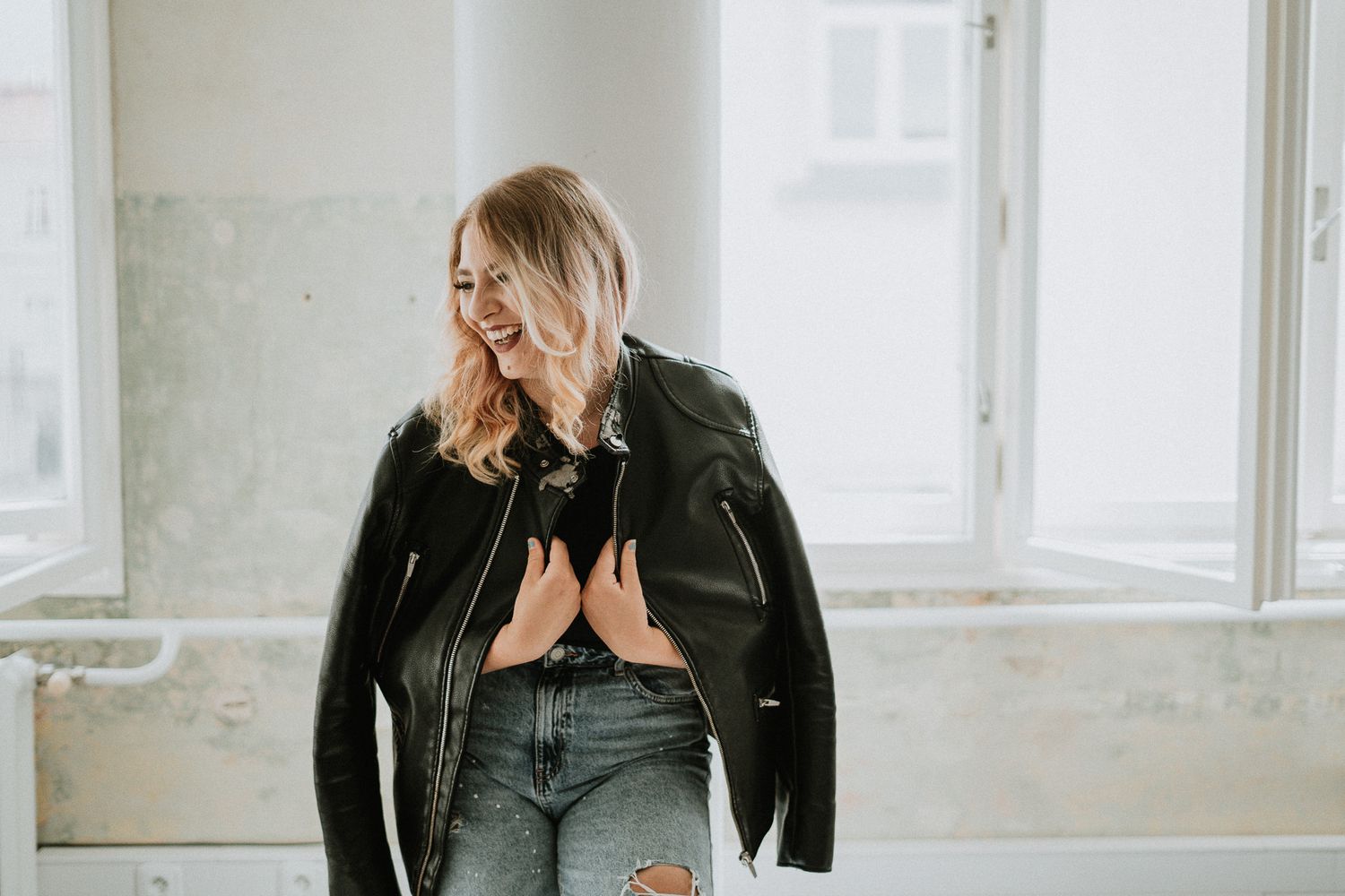 A person in a black leather jacket and ripped jeans poses casually by a window.