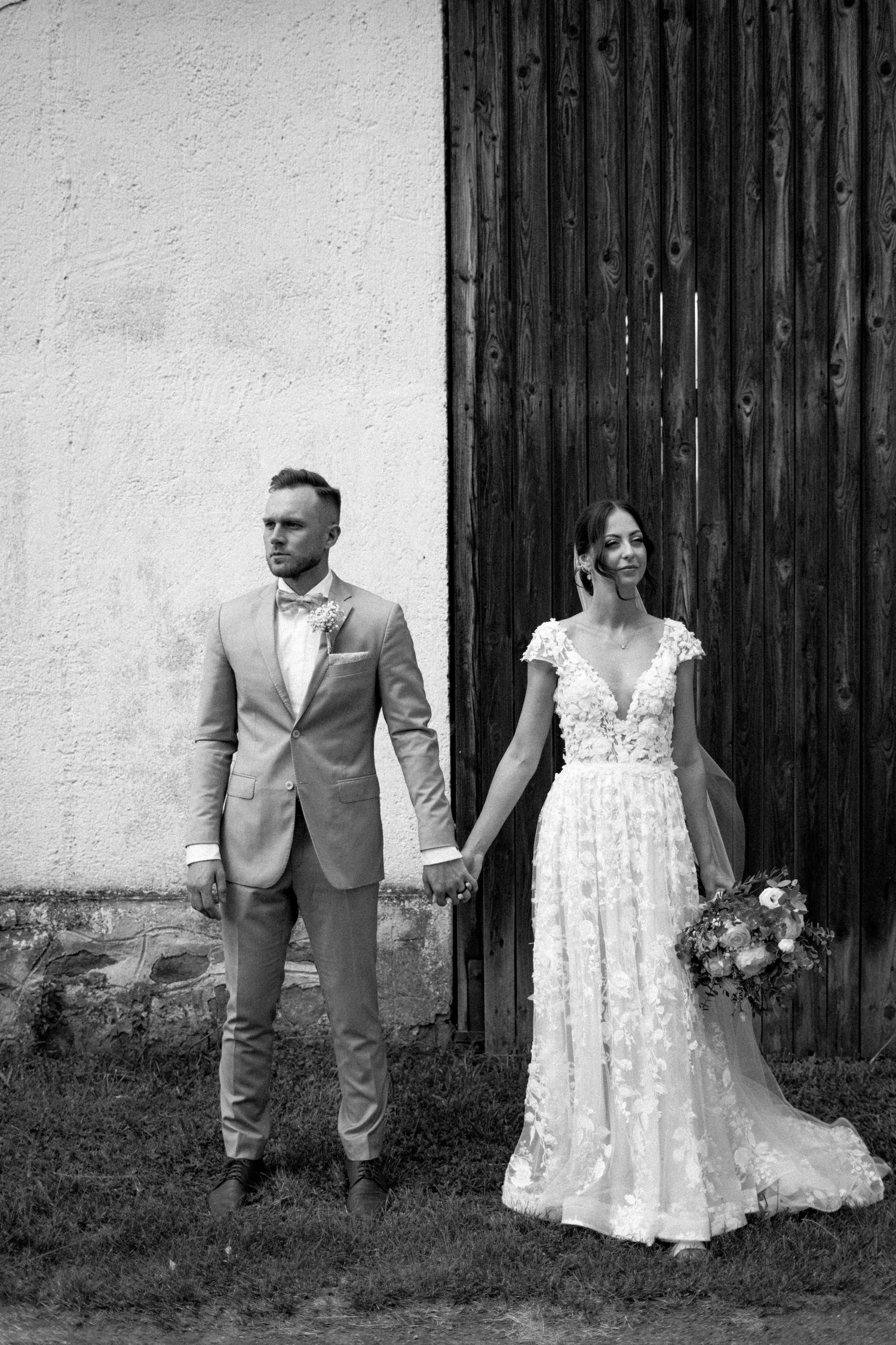 Elegant bride and groom in formal attire pose separately against rustic barn wall.