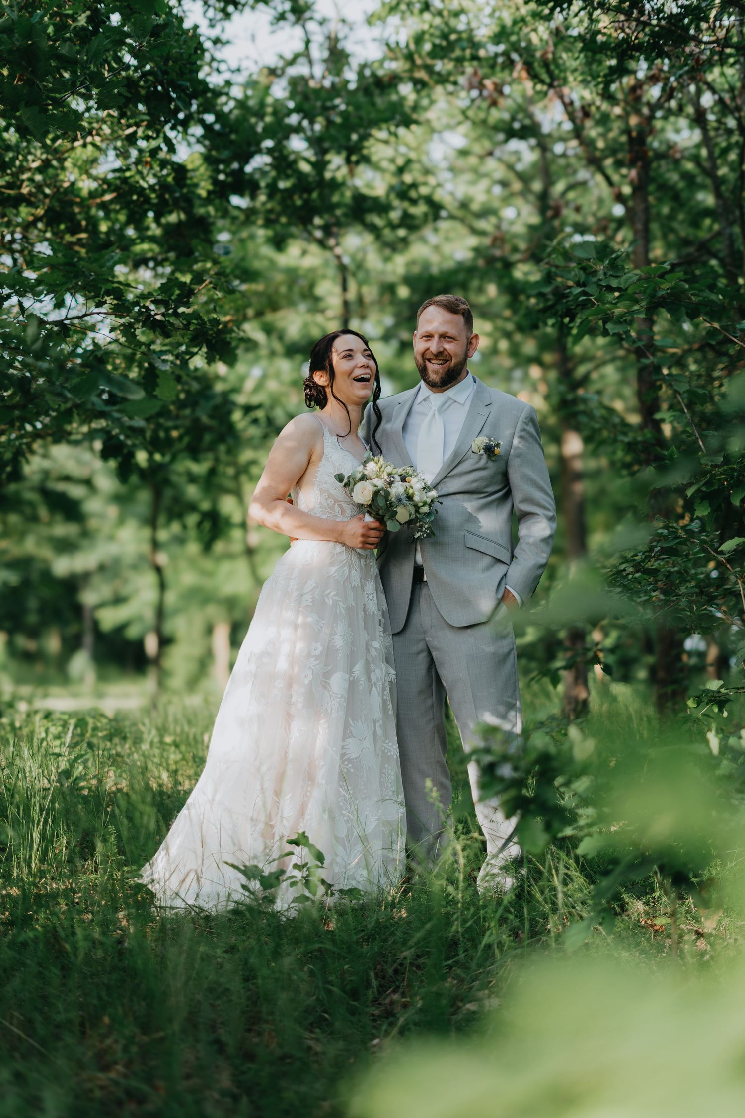 Beautiful outdoor wedding photo in a lush green forest setting, with a couple in grey suit and lace white gown.