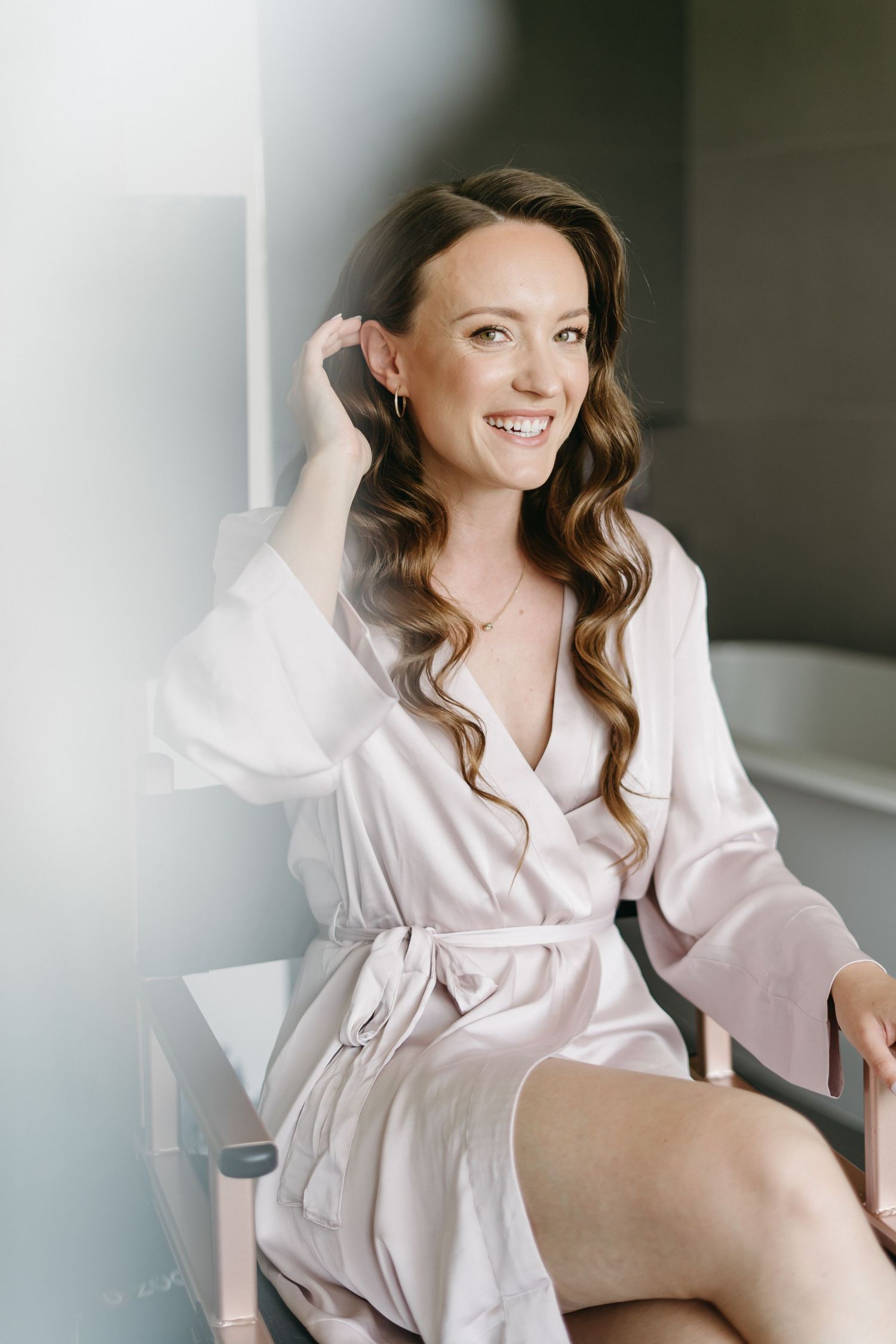 Woman in blush silk robe with long wavy hair smiles while getting ready on her wedding day.