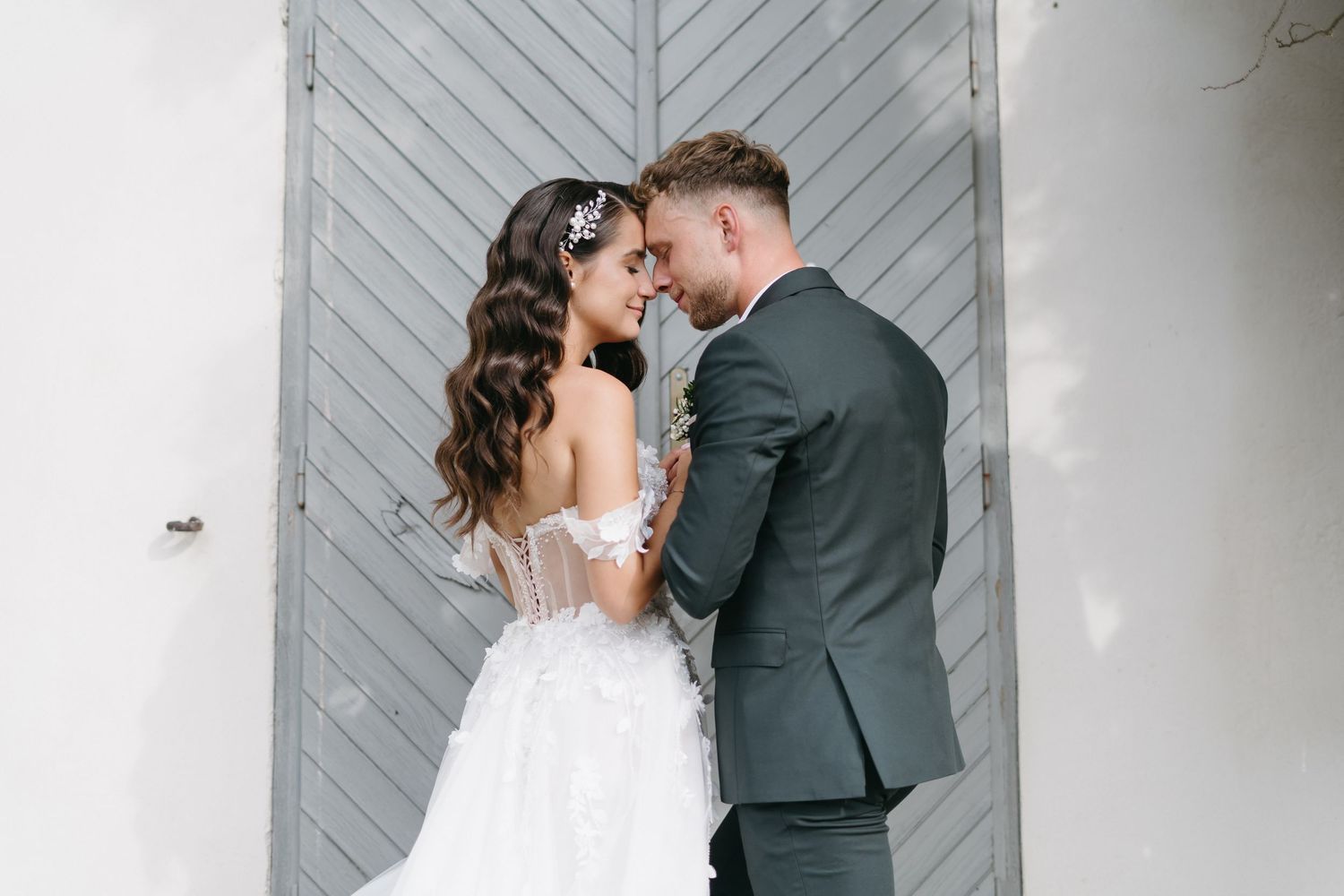 Bride and groom share a romantic moment in front of a rustic gray door, bride in lace gown with floral hairpiece.