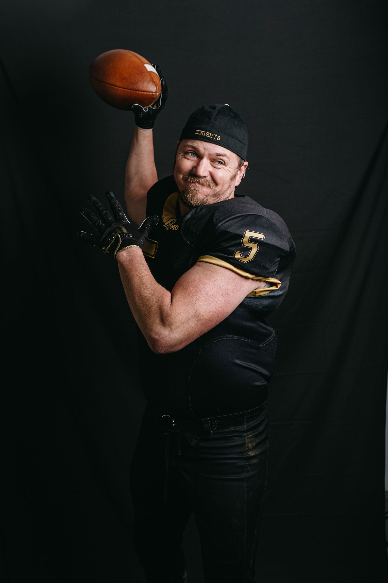 Athletic figure in black uniform with number 5 holding football overhead against dark background.