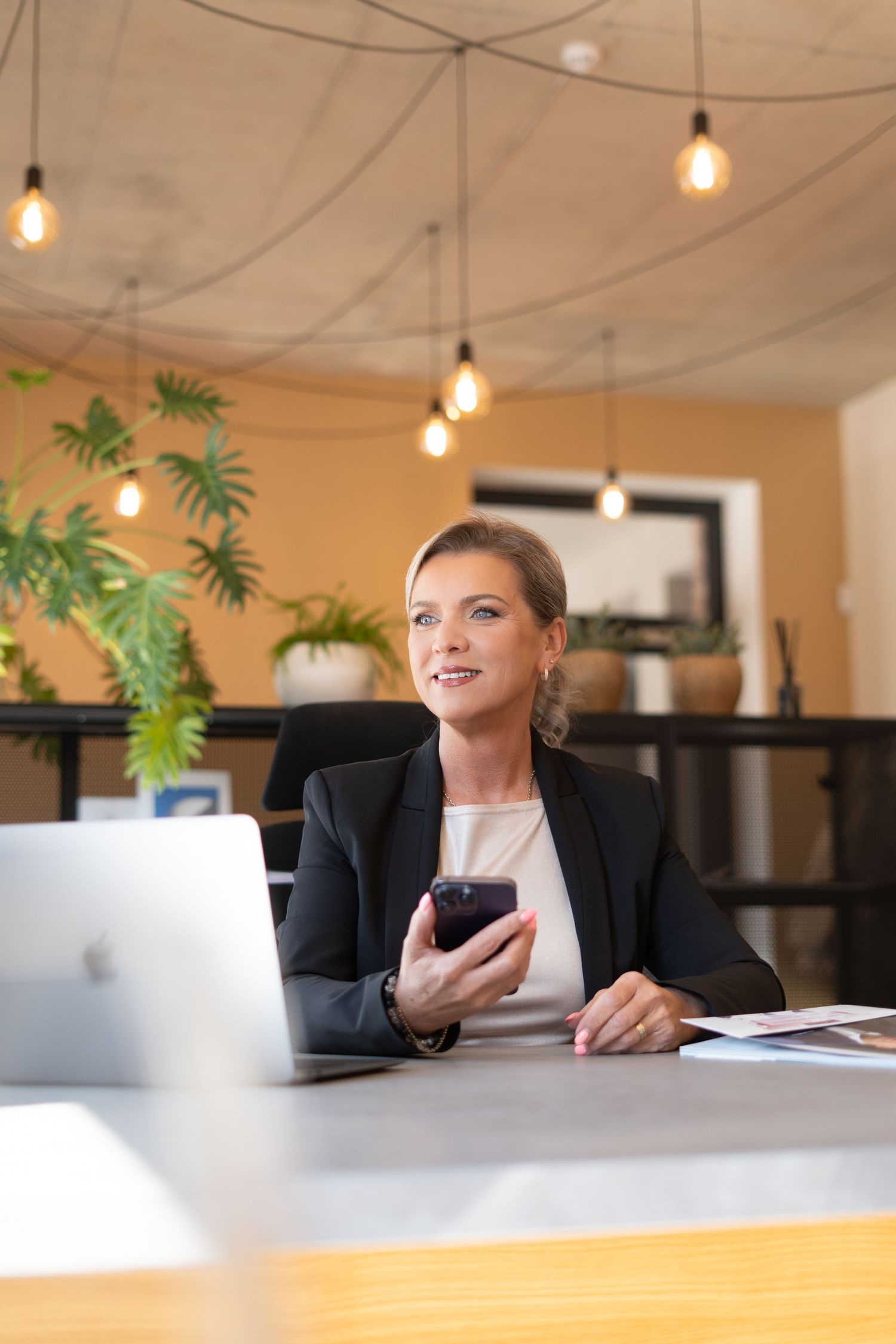 Professional working at desk with laptop and phone under hanging pendant lights in modern office space.