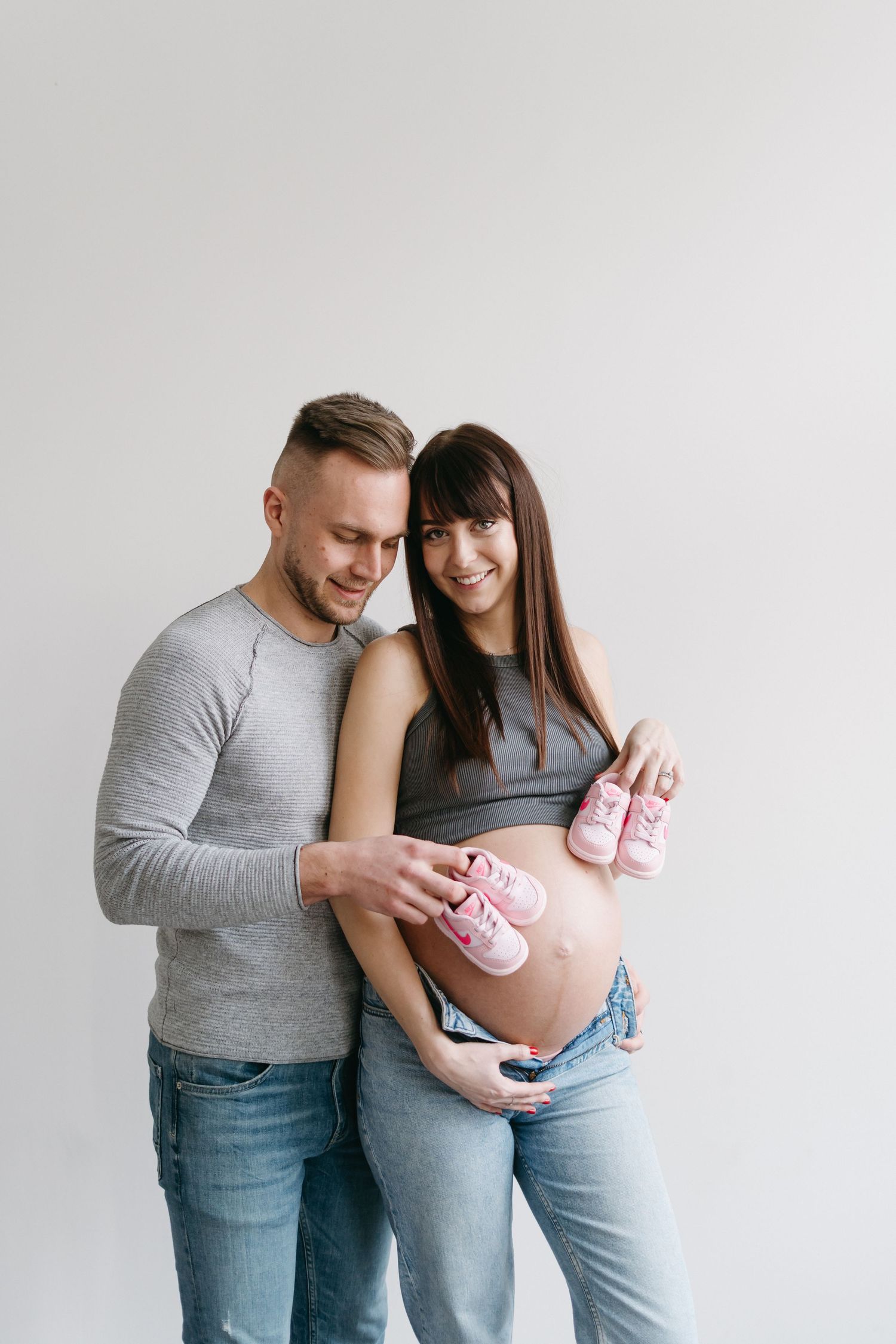 Happy couple holding pink baby shoes against pregnant belly in studio setting.