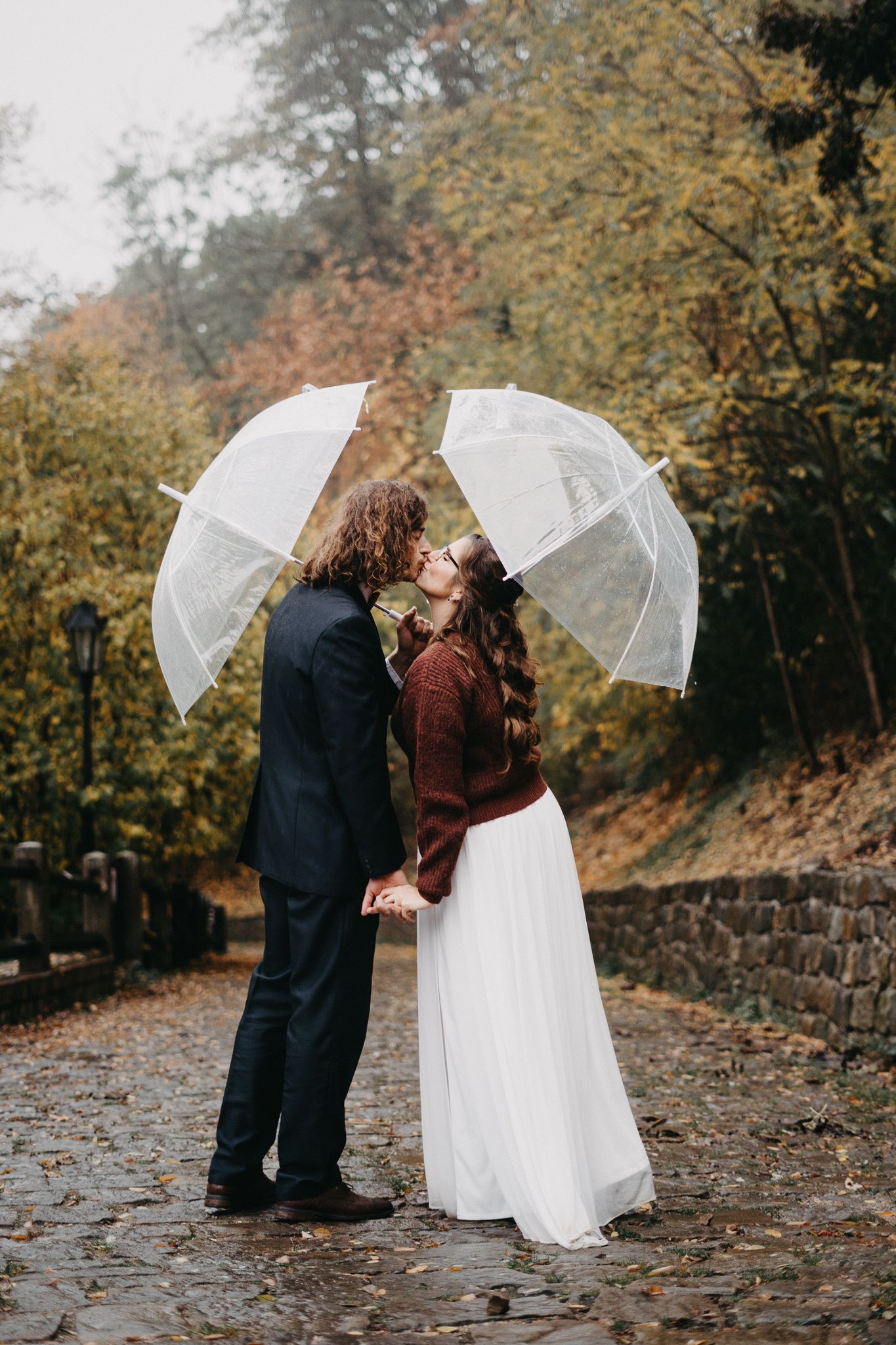 Two people share a romantic autumn moment under clear umbrellas on a stone path surrounded by fall foliage.