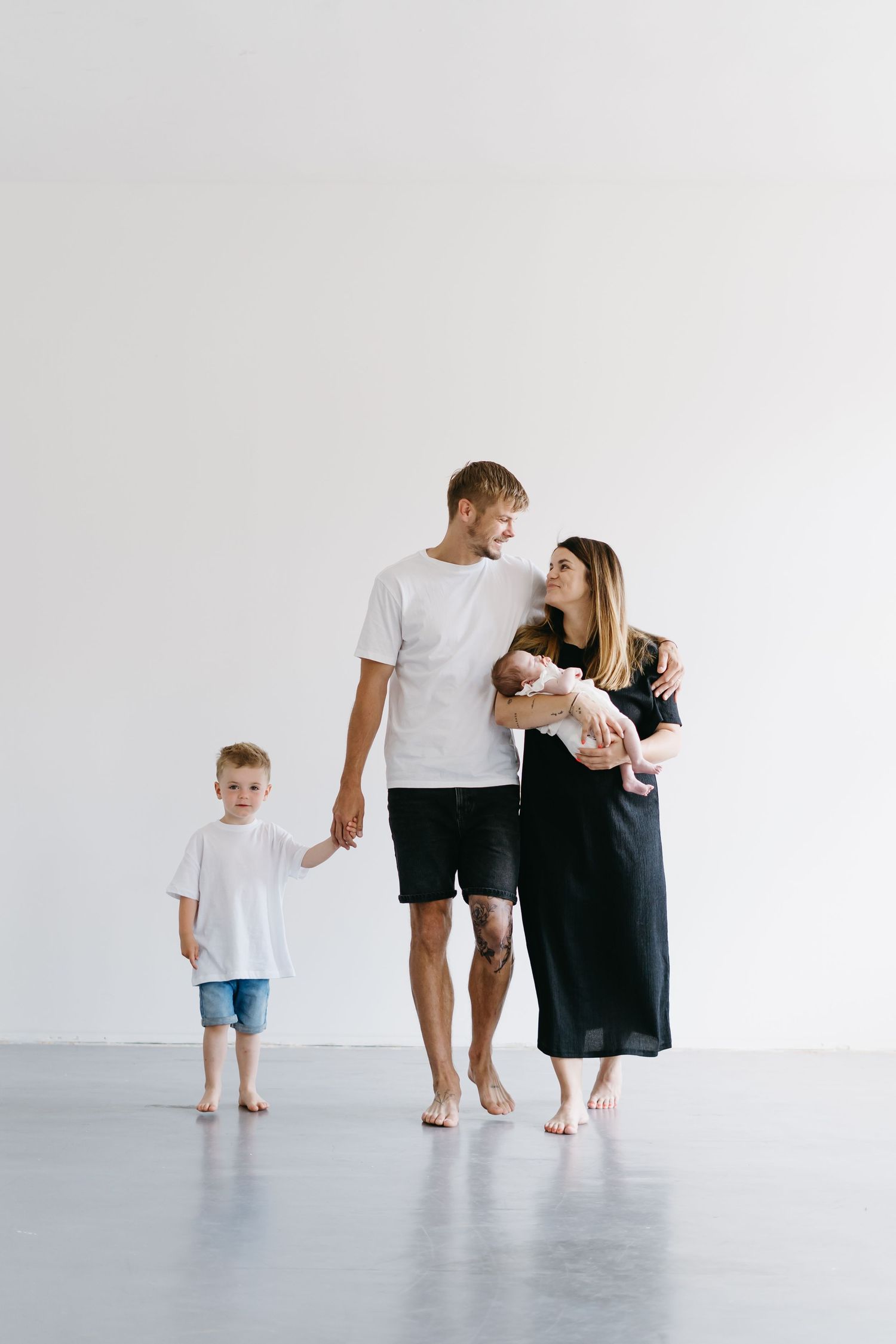 A family of three stands together against a white wall in matching white outfits.