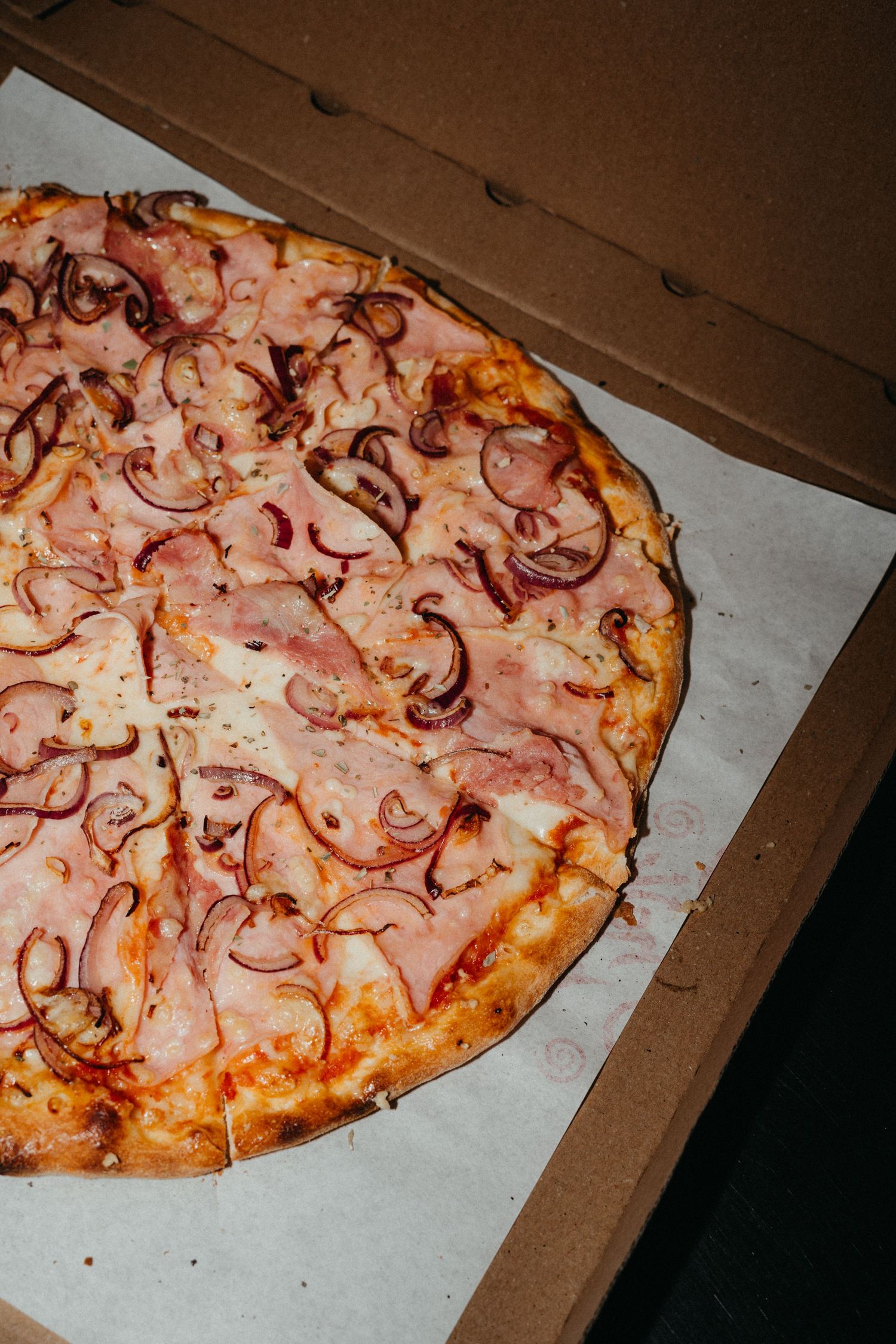 A pizza topped with thinly sliced red onions and ham sits in a cardboard delivery box on white paper.