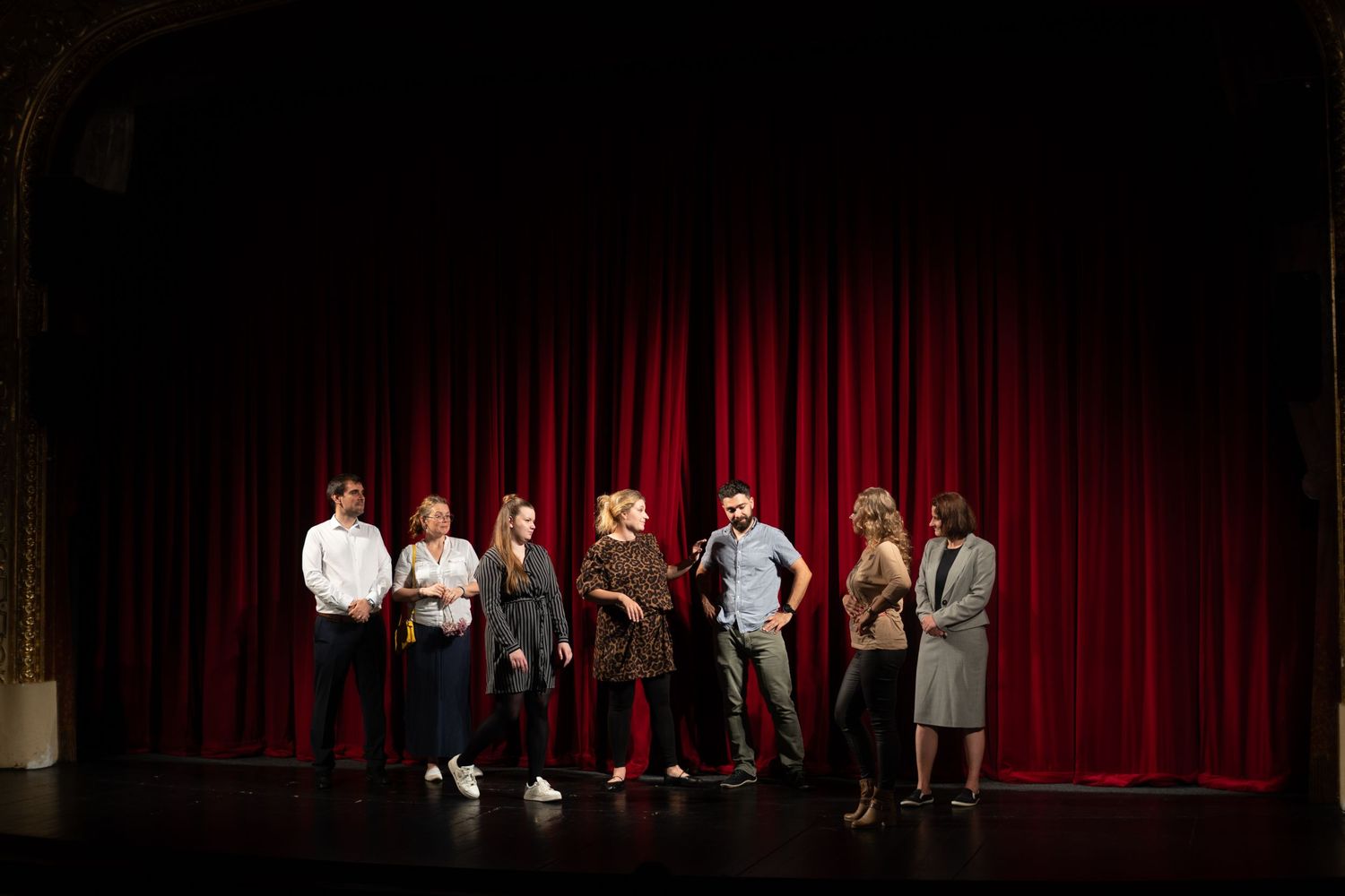 Performers stand on a stage in front of a red velvet curtain during a theater production.