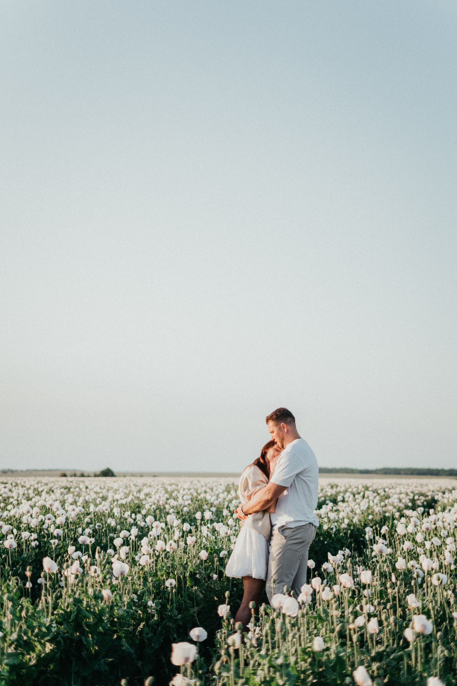 A couple shares intimate moments in a dreamy white poppy field stretching to the horizon under an overcast sky.