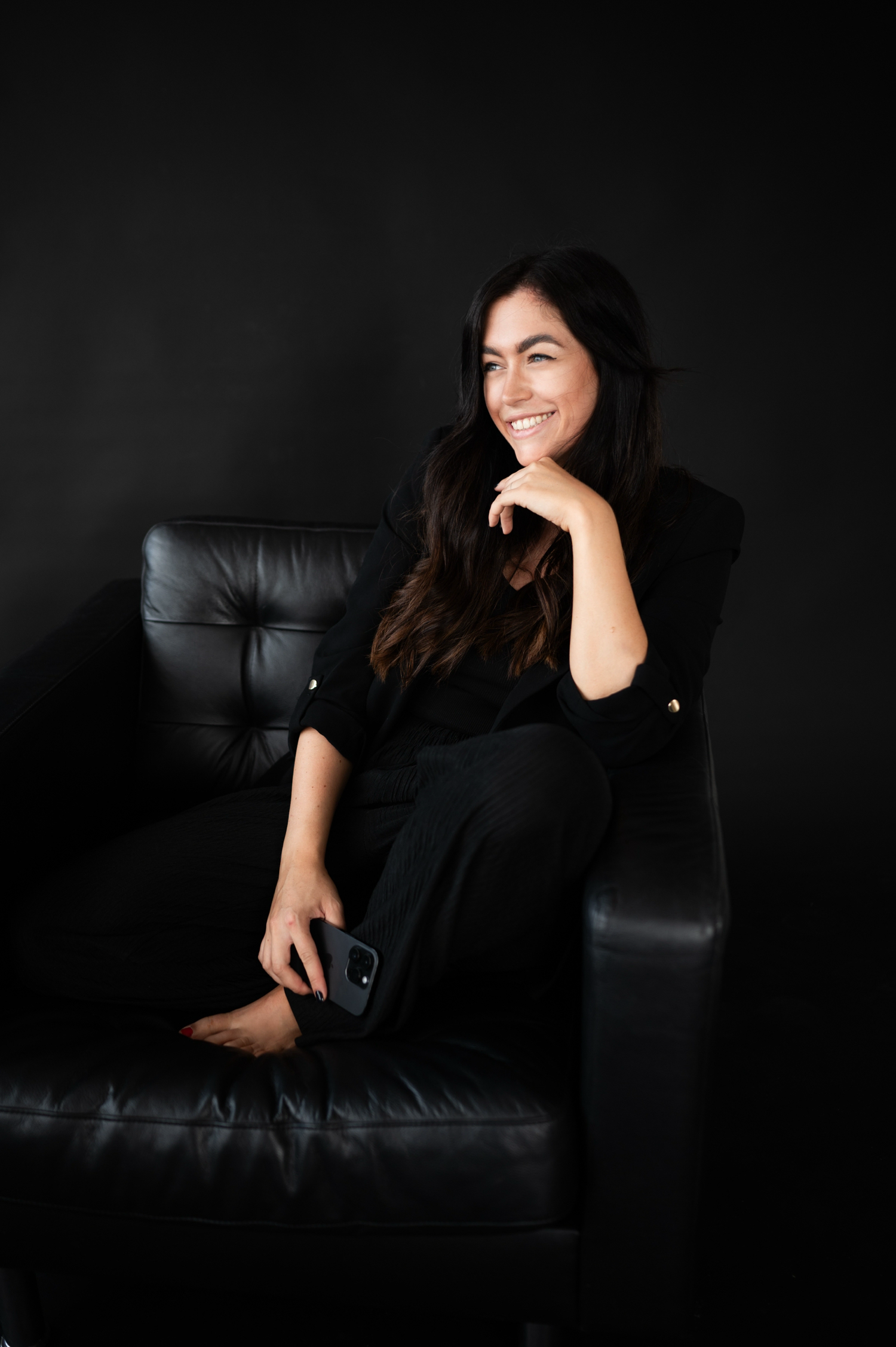 Figure in black clothing reclines on a dark leather couch against a moody black background in artistic portrait series.