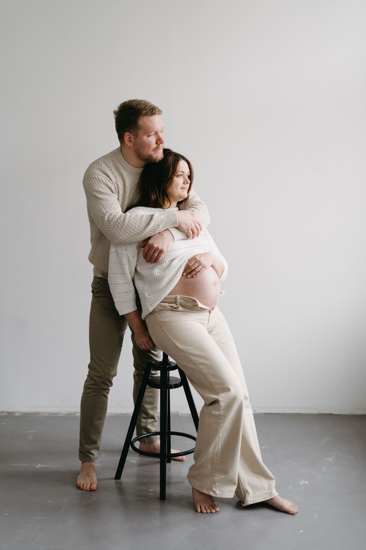 A couple poses with a bar stool during their indoor maternity photoshoot in neutral colors.